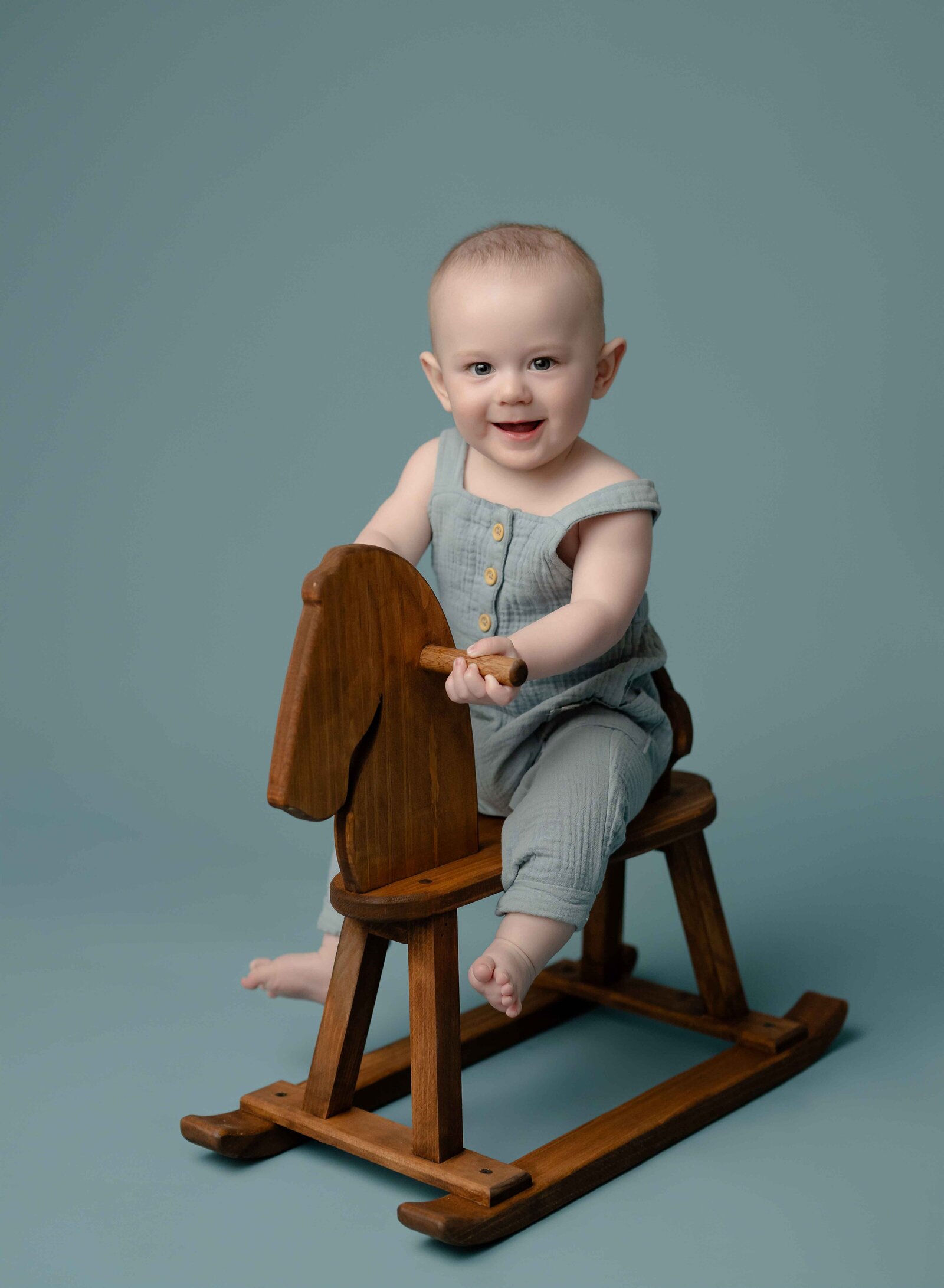 Baby in blue overalls sitting on wooden horse and smiling. Photo taken at Atlanta Baby Photo Studio.