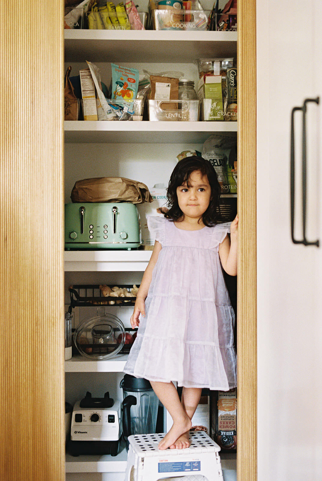 A candid image of a young girl standing confidently in an organized pantry, dressed in a flowing white dress and framed by the open pantry doors.
