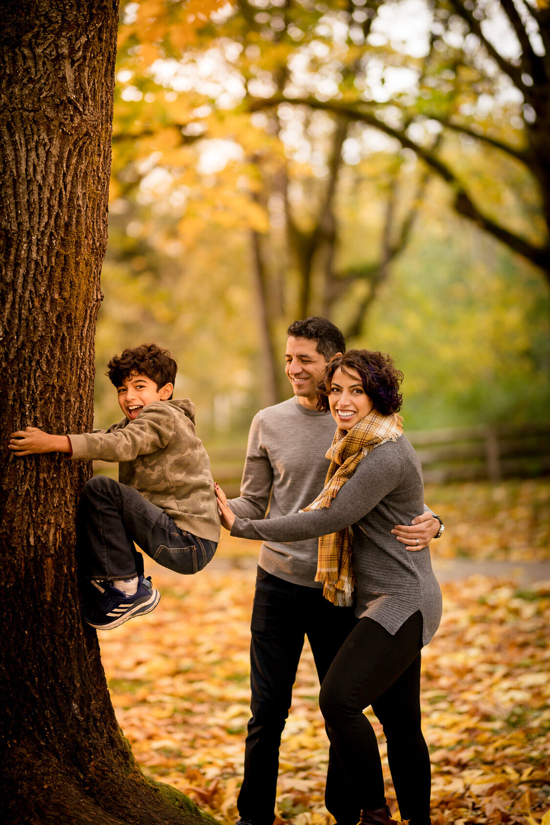Fall family portrait in Redmond, Washington of a joyful boy climbing a tree, with his smiling parents behind him. This candid image captures authentic family moments in Maë Lans Photography’s signature golden, glowy style.