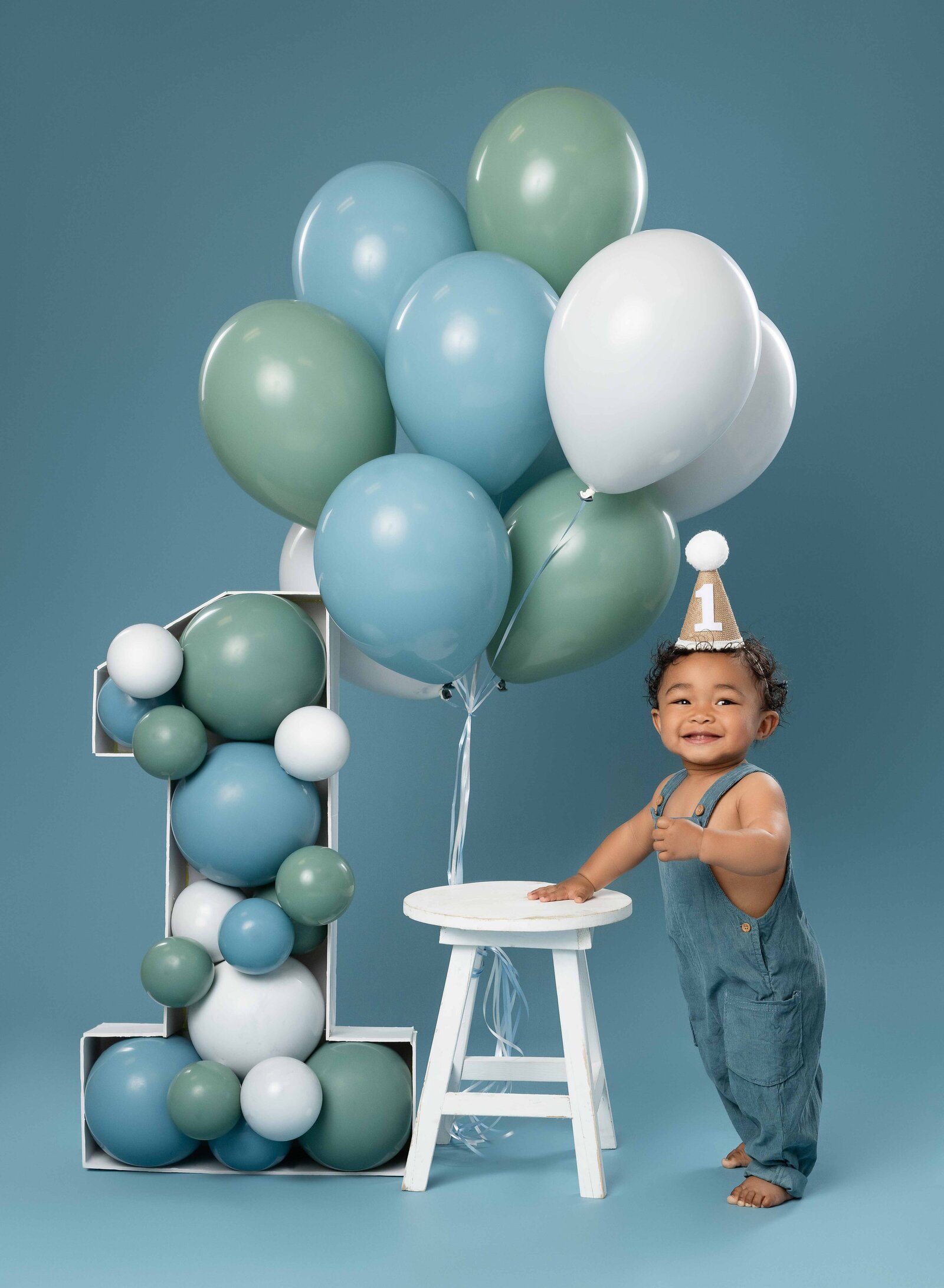 1 year old taking birthday photos, staning next to a stool with balloons tied to it and a balloon filled marquee, smiling.