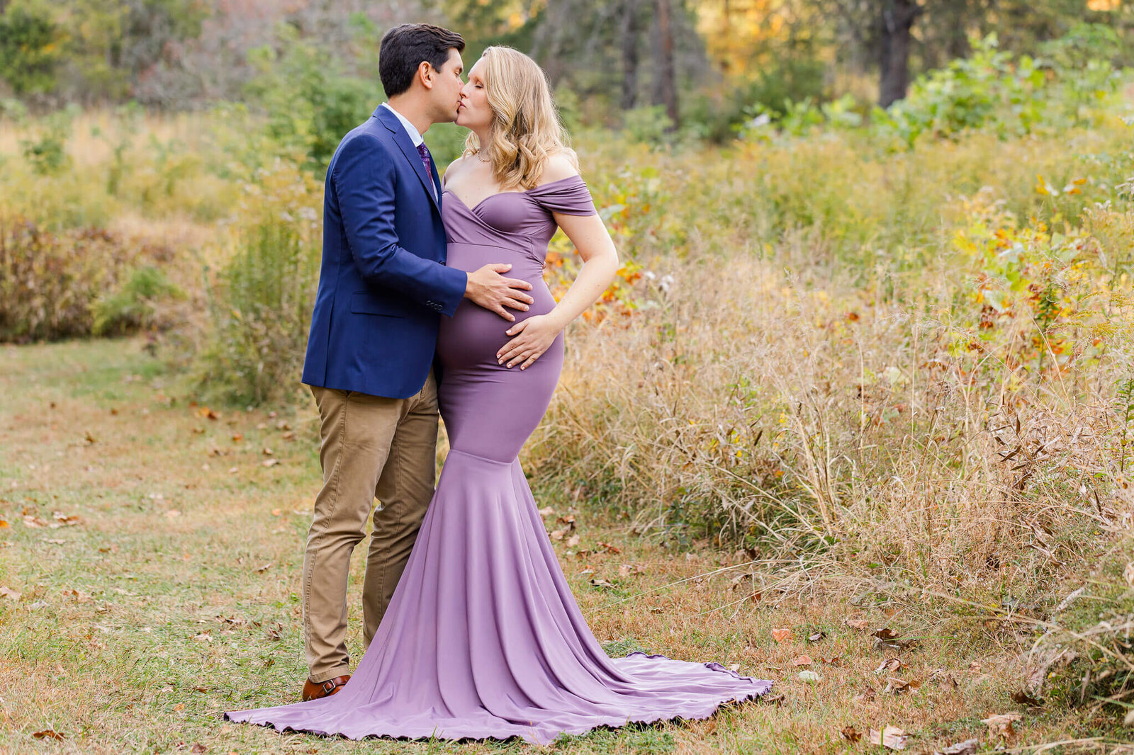 A pregnant woman and her husband embracing and kissing in a field at sunset.