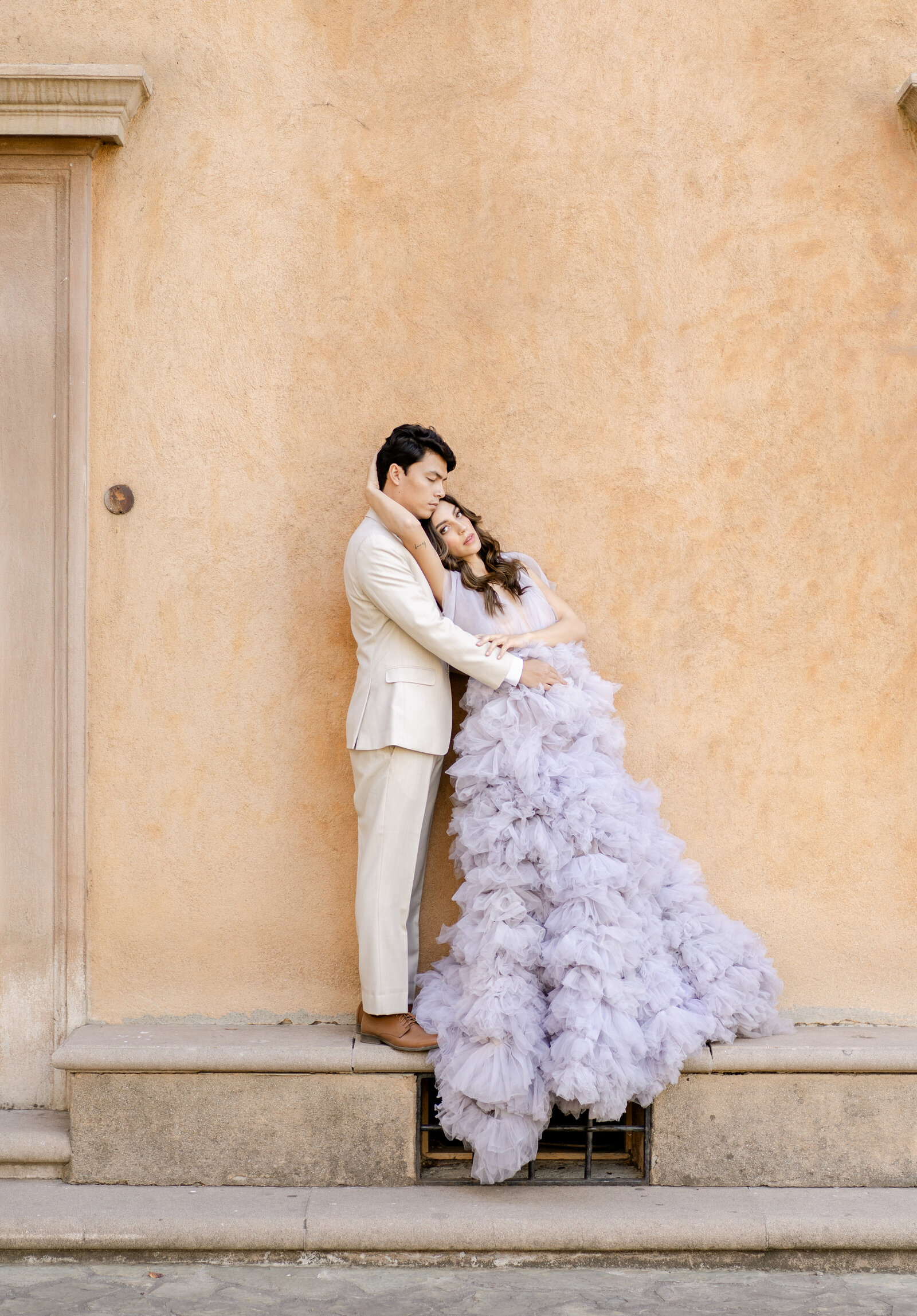 Portrait of a bride in a lavender dress standing with a groom in an ivory suit near a peach-colored building.