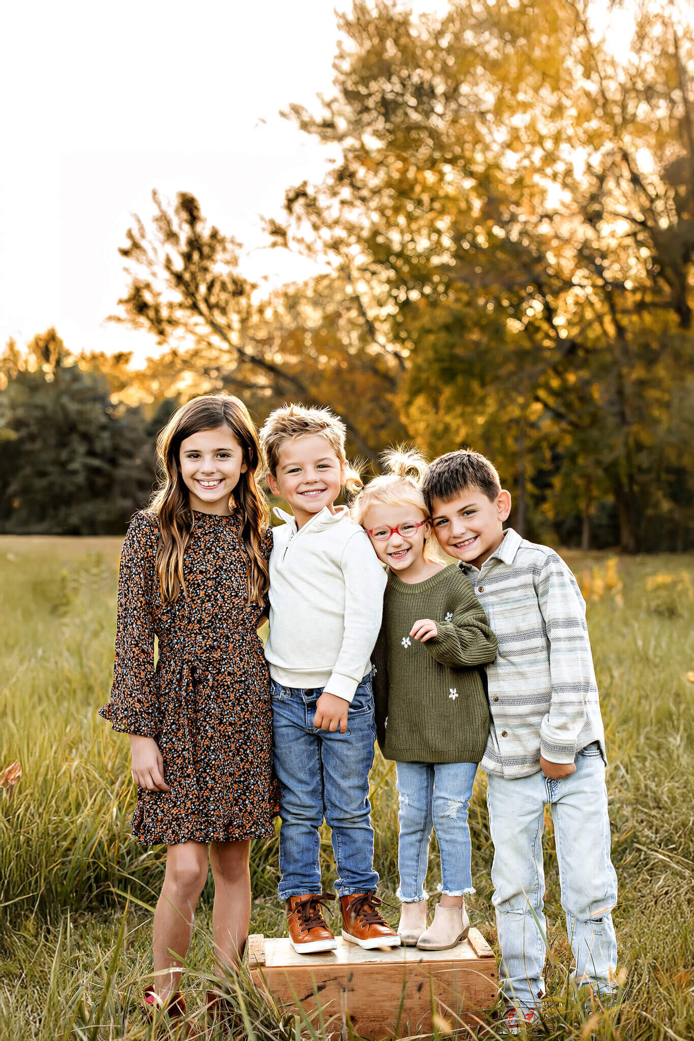 Four children stand closely together in a grassy field during sunset, smiling with autumn trees in the background. Captured like outdoor portraits, one child stands on a wooden crate, casually dressed in fall attire.