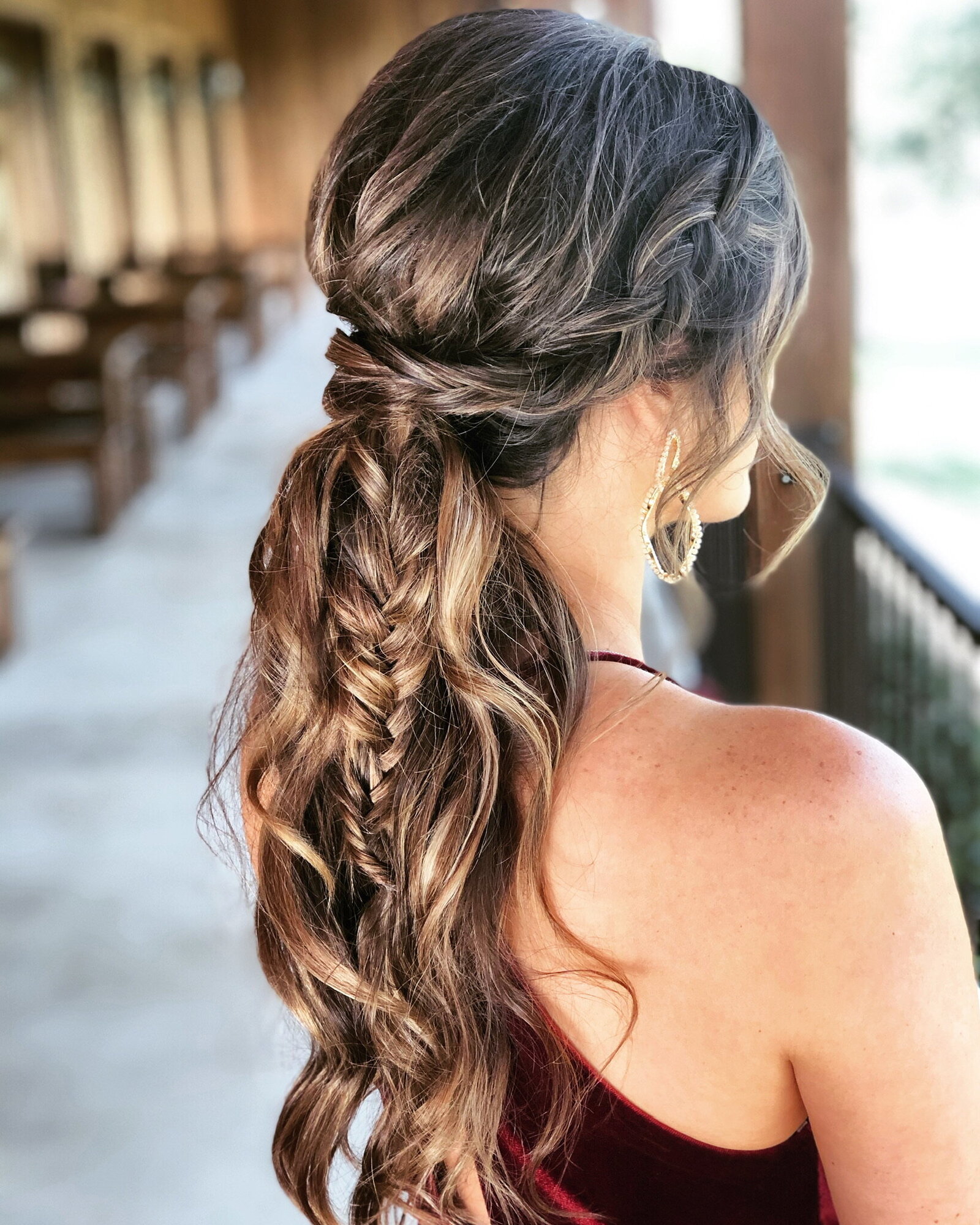 Woman with long, braided hairstyle wearing a maroon dress, standing on a porch with wooden railings. Experience true bridal beauty in Texas as her look embodies elegance and charm.