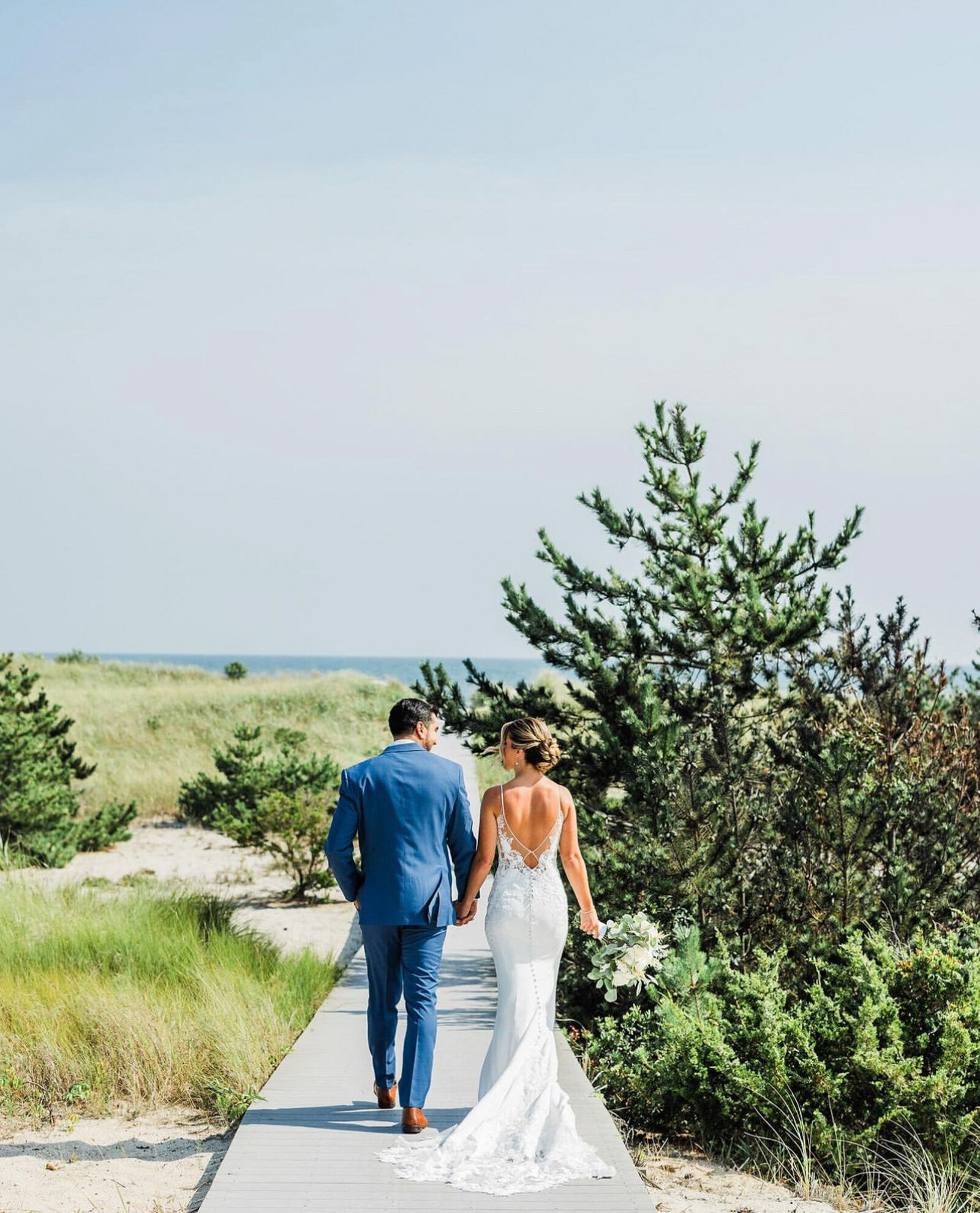 bride and groom walk hand in hand on a wooden boardwalk surrounded by greenery, heading towards the beach