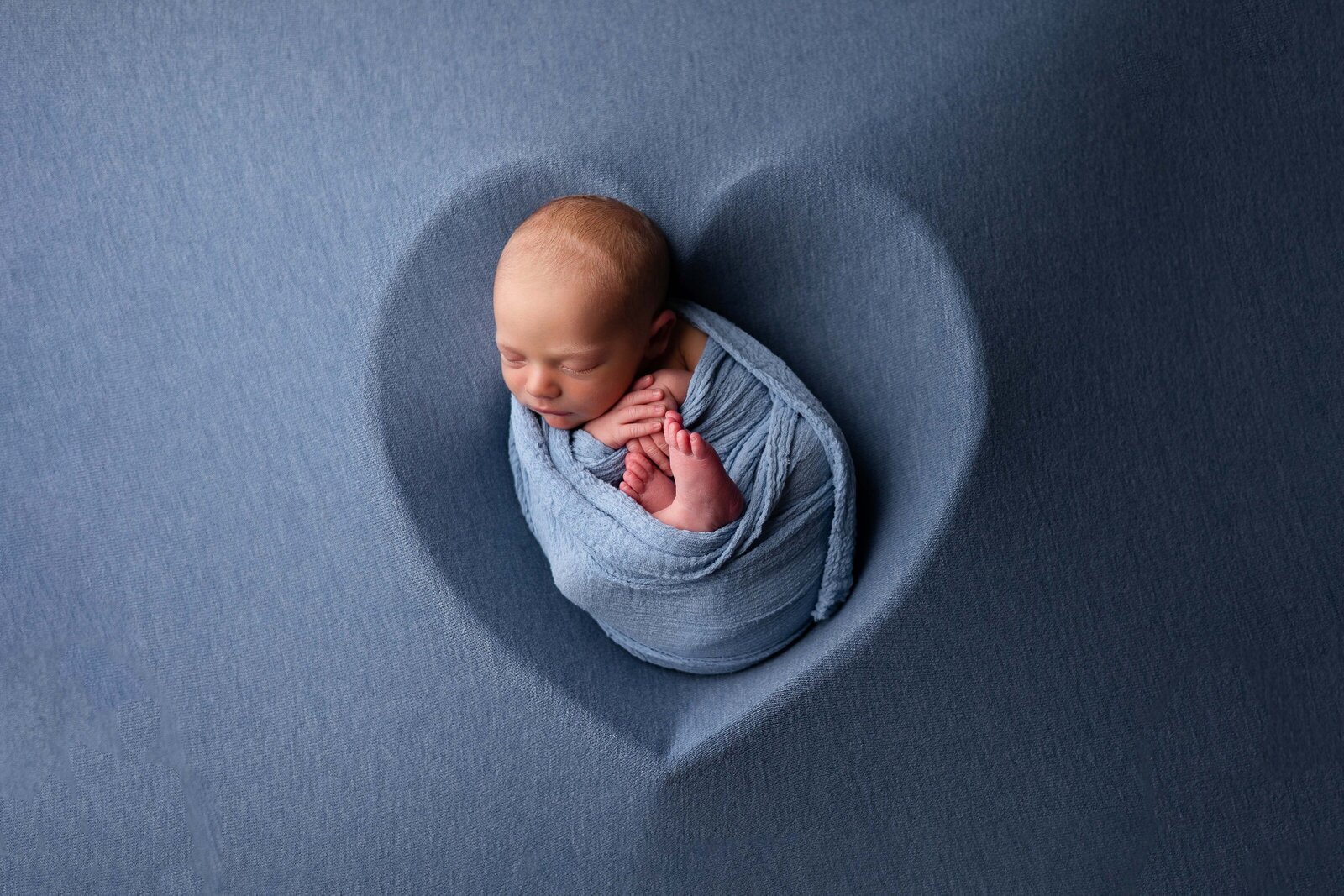 Newborn baby boy wrapped in blue laying in heart shaped bowl