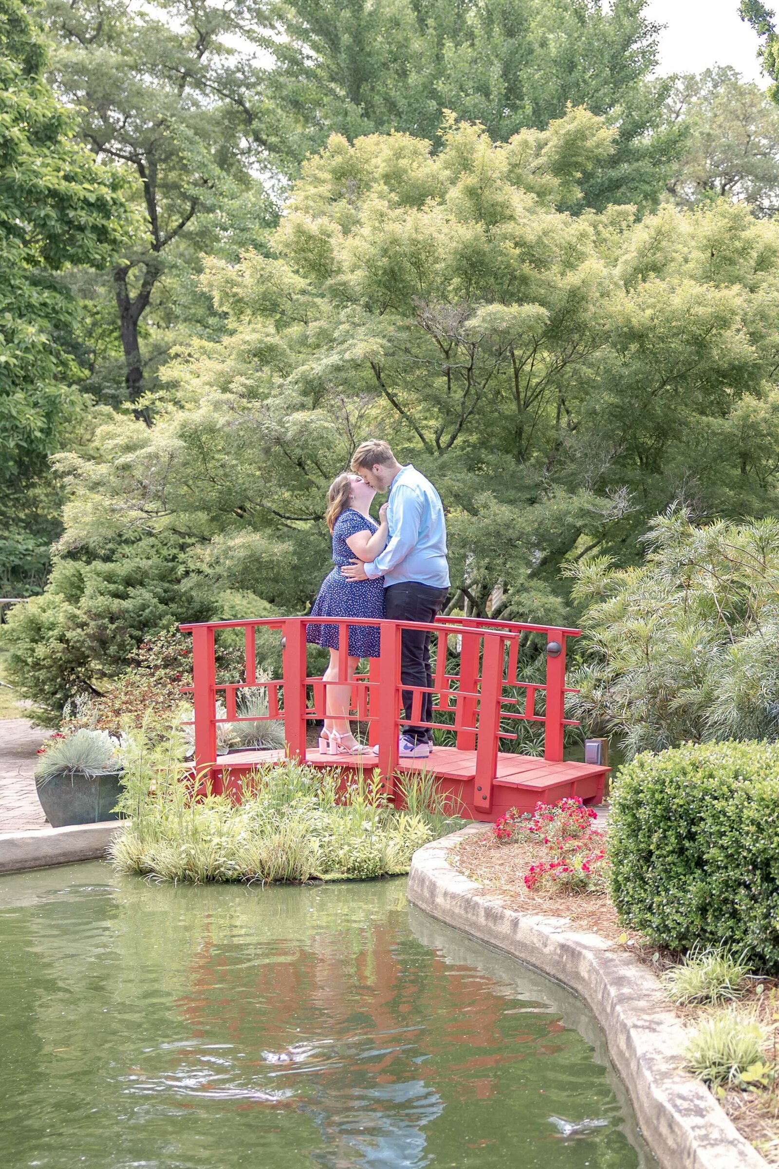 A romantic couple shares a tender moment on a bright red bridge surrounded by lush greenery and a tranquil pond. The man leans in to kiss the woman, who wears a blue dress. This charming scene reflects the elegance and serenity often captured in North Carolina engagement photography.  