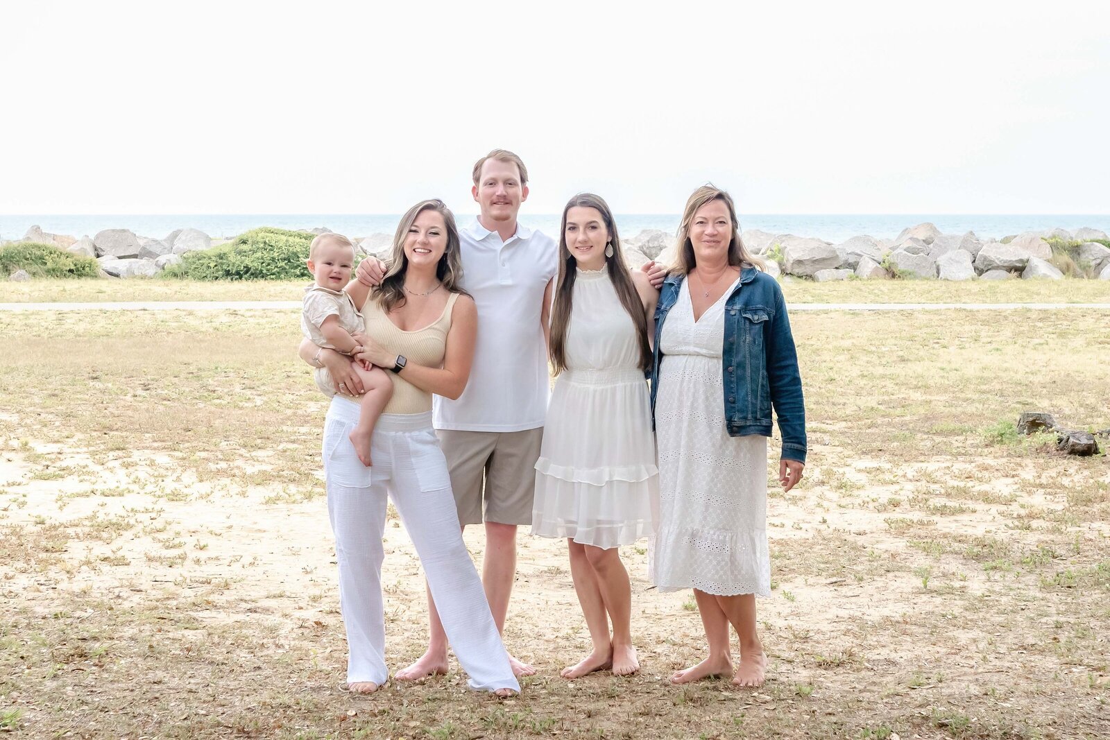  A family poses together near a rocky coastline, with the ocean visible in the background. Dressed in white and denim, they exude a relaxed, coastal vibe. This image is ideal for professional family photography in NC, capturing timeless beachside moments.