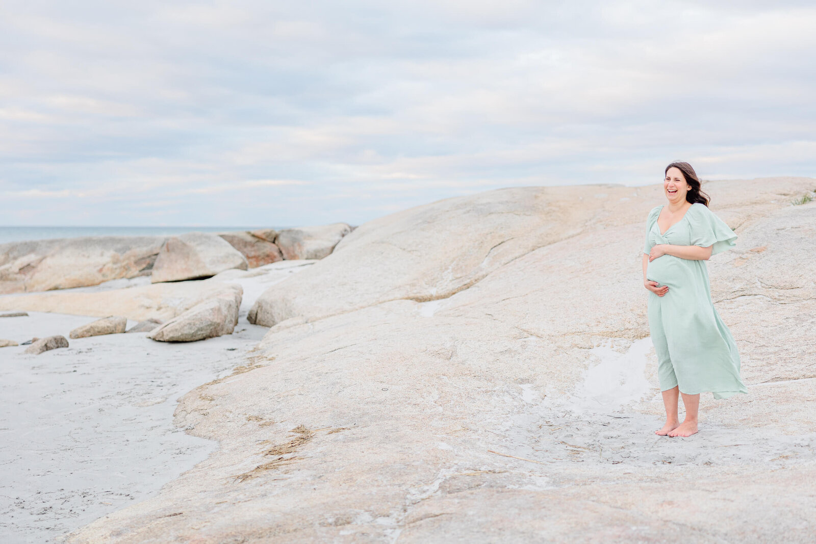 Pulled back view of a pregnant woman holding her baby bump and laughing in a rocky area of a Northshore Boston beach