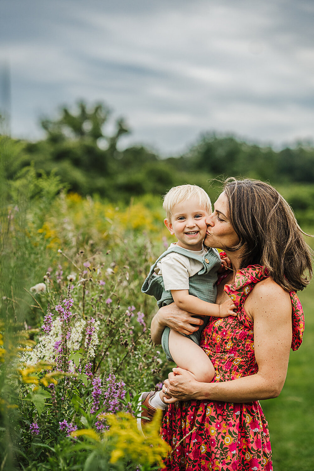 mom in floral dress kisses toddler boy among wildflowers