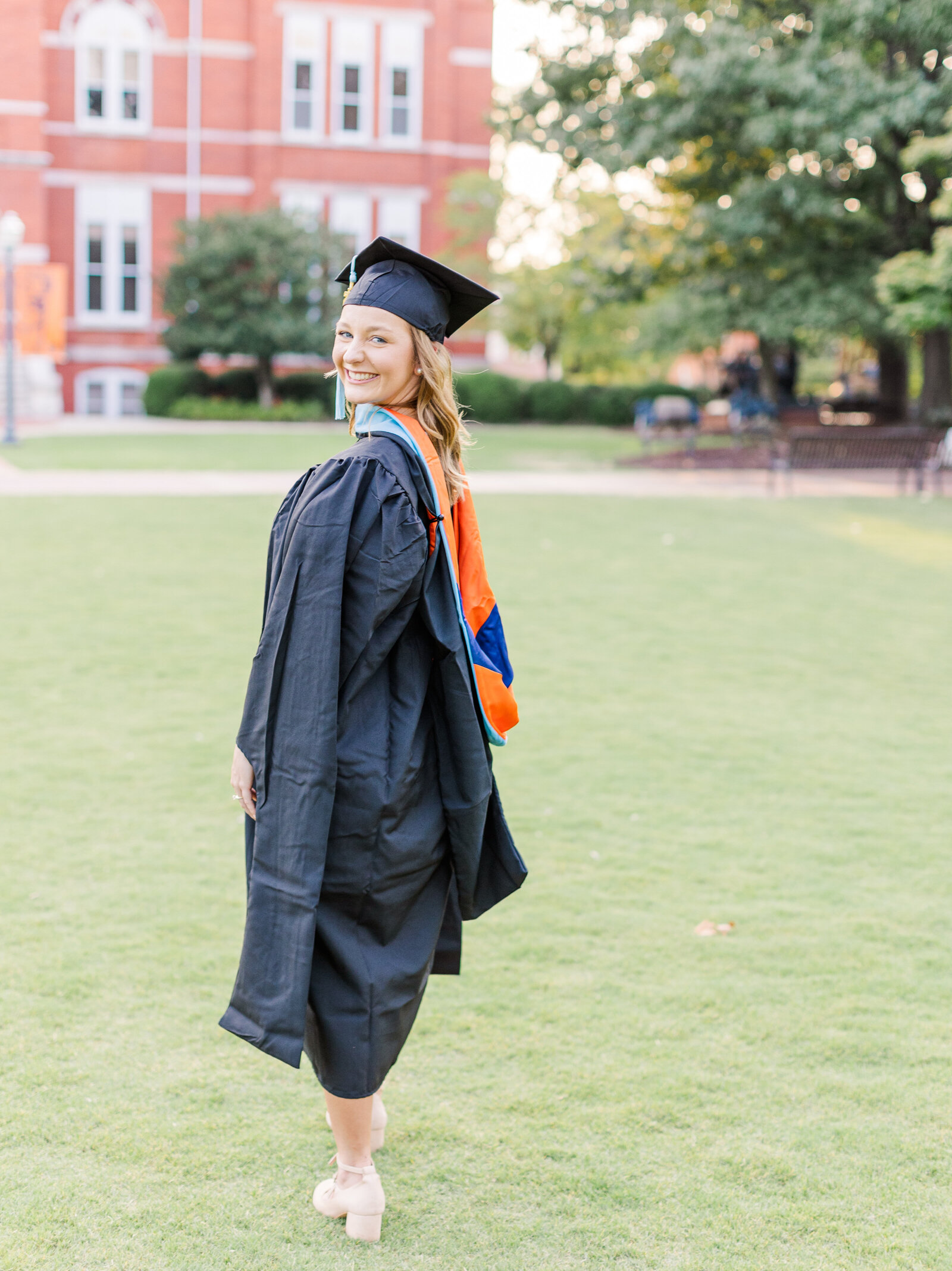 Shelby showing off her graduation gown and cap with a joyful smile, photographed at a lush green campus by Abby Bell Photography.