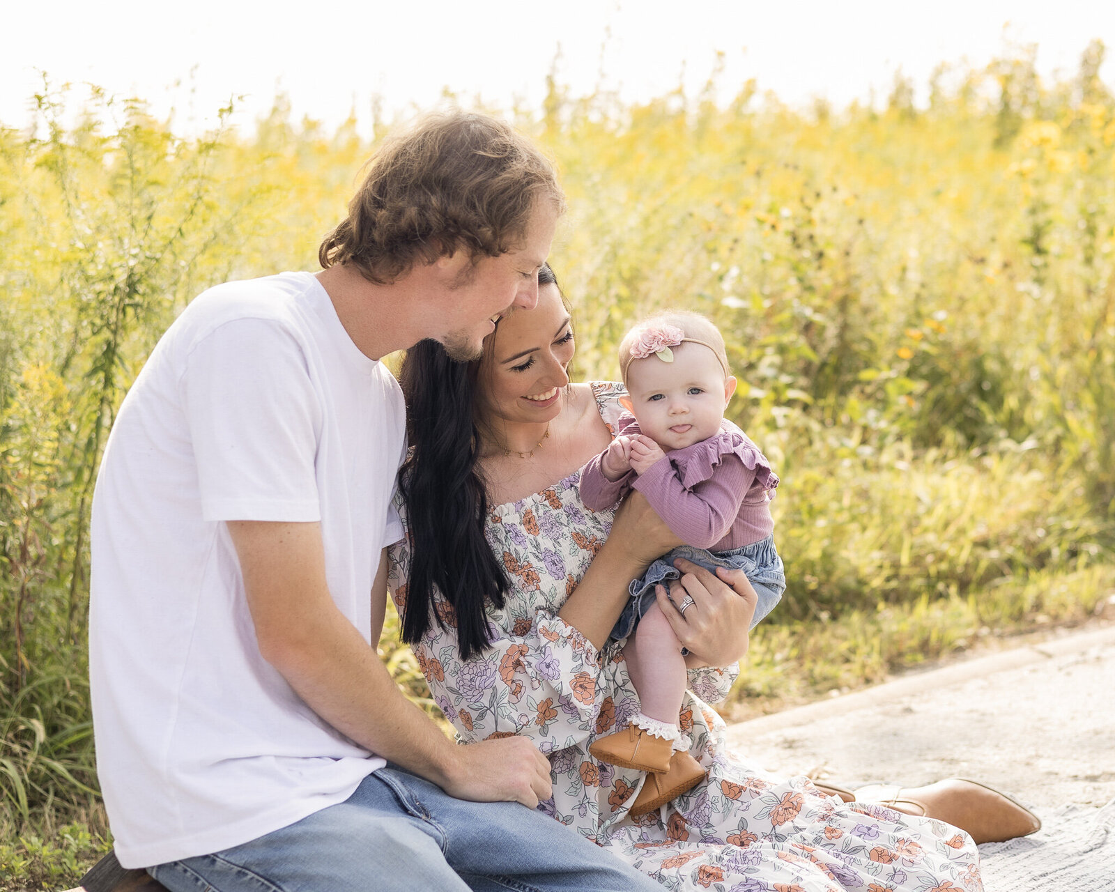 A family of 3 sitting on the ground at a wildlife sanctuary.  Father has brown blonde hair wearing a white shirt, blue jeans, looking and smiling at their 6 mo old daughter, Mom has black long hair, wearing a white dress with flower print, and tan boots, smiling and looking at their 6 month old daughter.  The 6 month daughter is wearing a pink headband with flowers, wearing a purple long sleet shirt, with a blue jean skirt and brown shoes, looking at the camera.