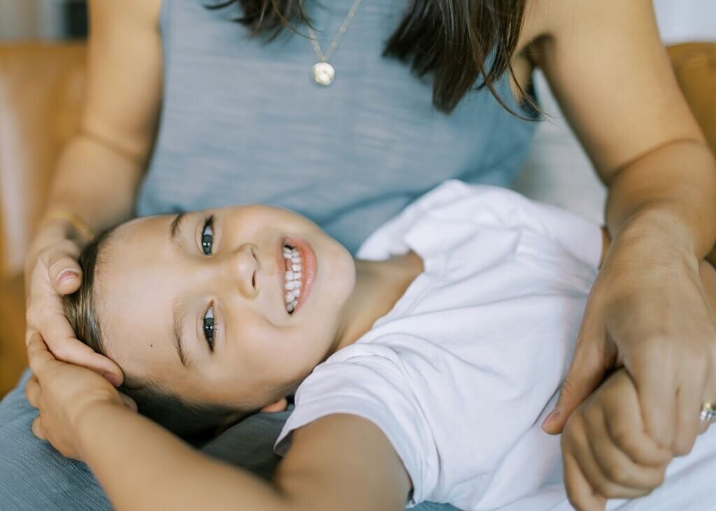 Young boy smiling at the camera wild holding hands and laying in his mother's lap.
