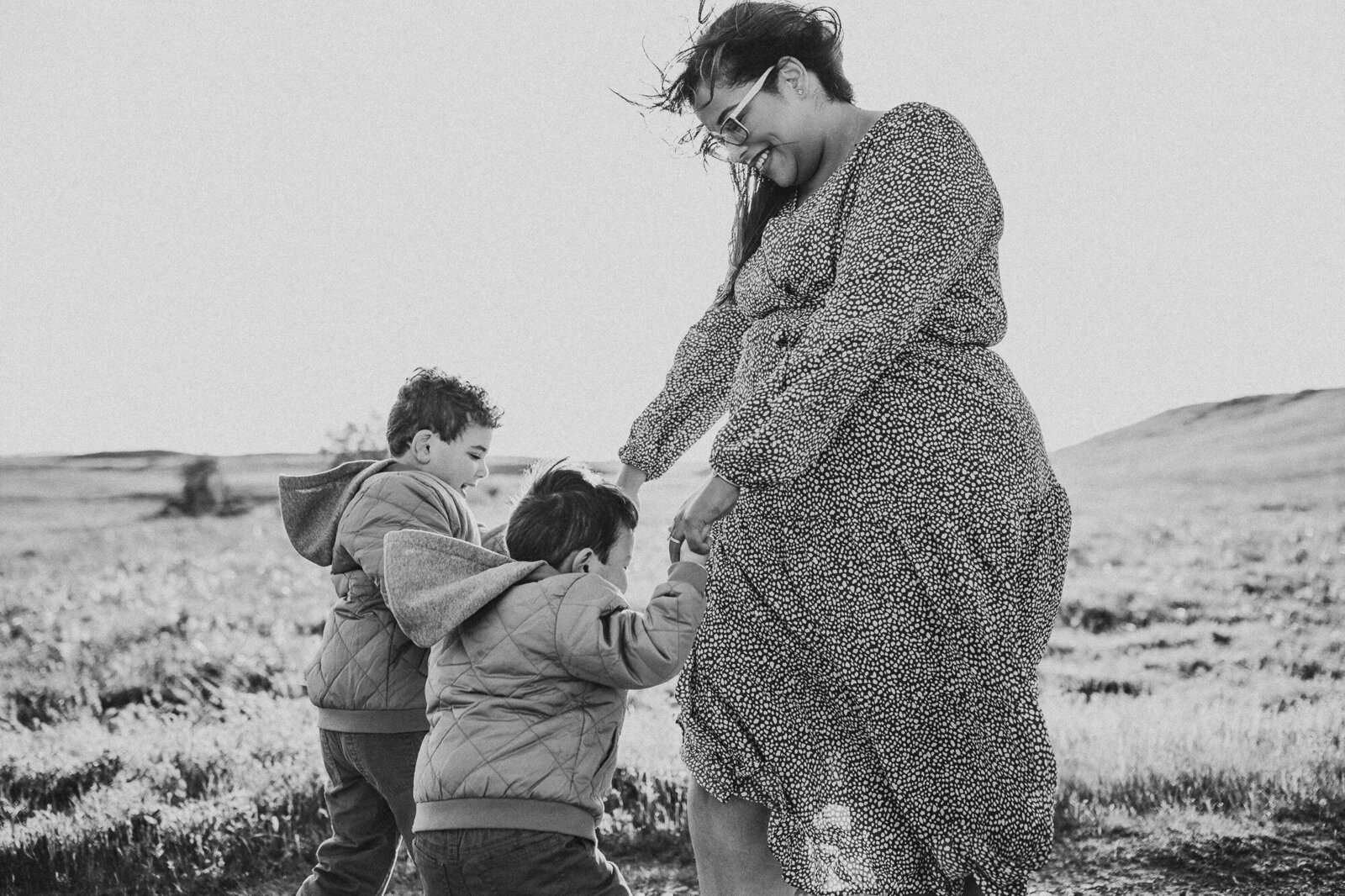 Mother and two young sons playing Ring Around the Rosie in a field in Oregon with the sun bursting behind them.