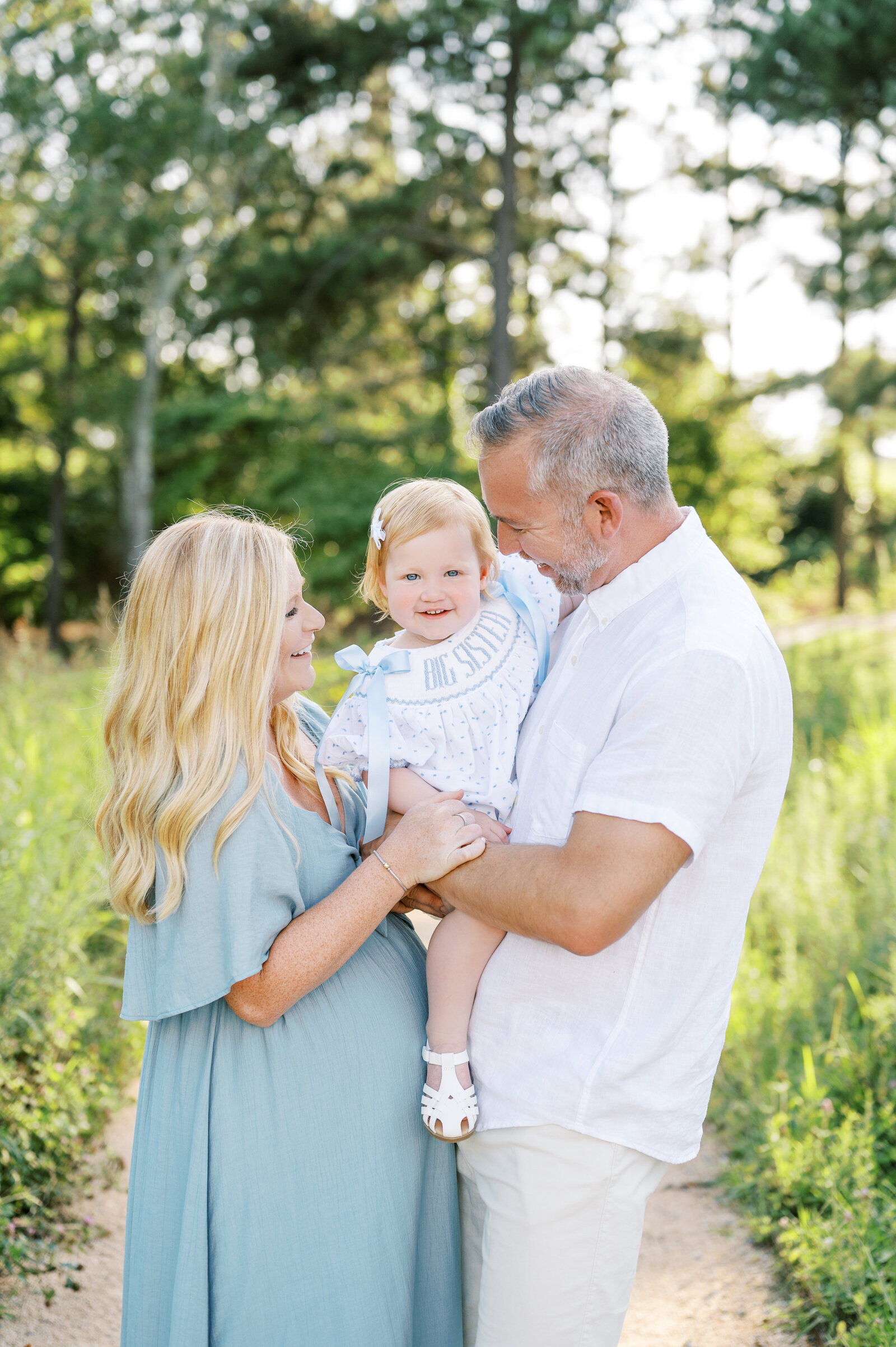 Pregnant mom in blue green dress and Dad in white shirt hold red-headed toddler girl in 'big sister' dress during family maternity photos at a park in Raleigh North Carolina