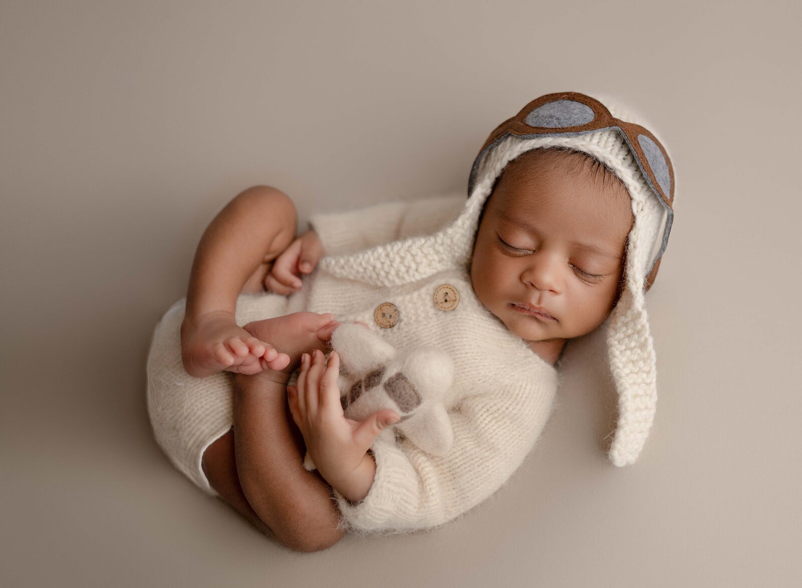 Baby boy sleeping on his back, holding a felted airplane and wearing an aviator bonnet. Newborn Photography session photographed at Jenni Nikki's photo studio in Marietta, GA.