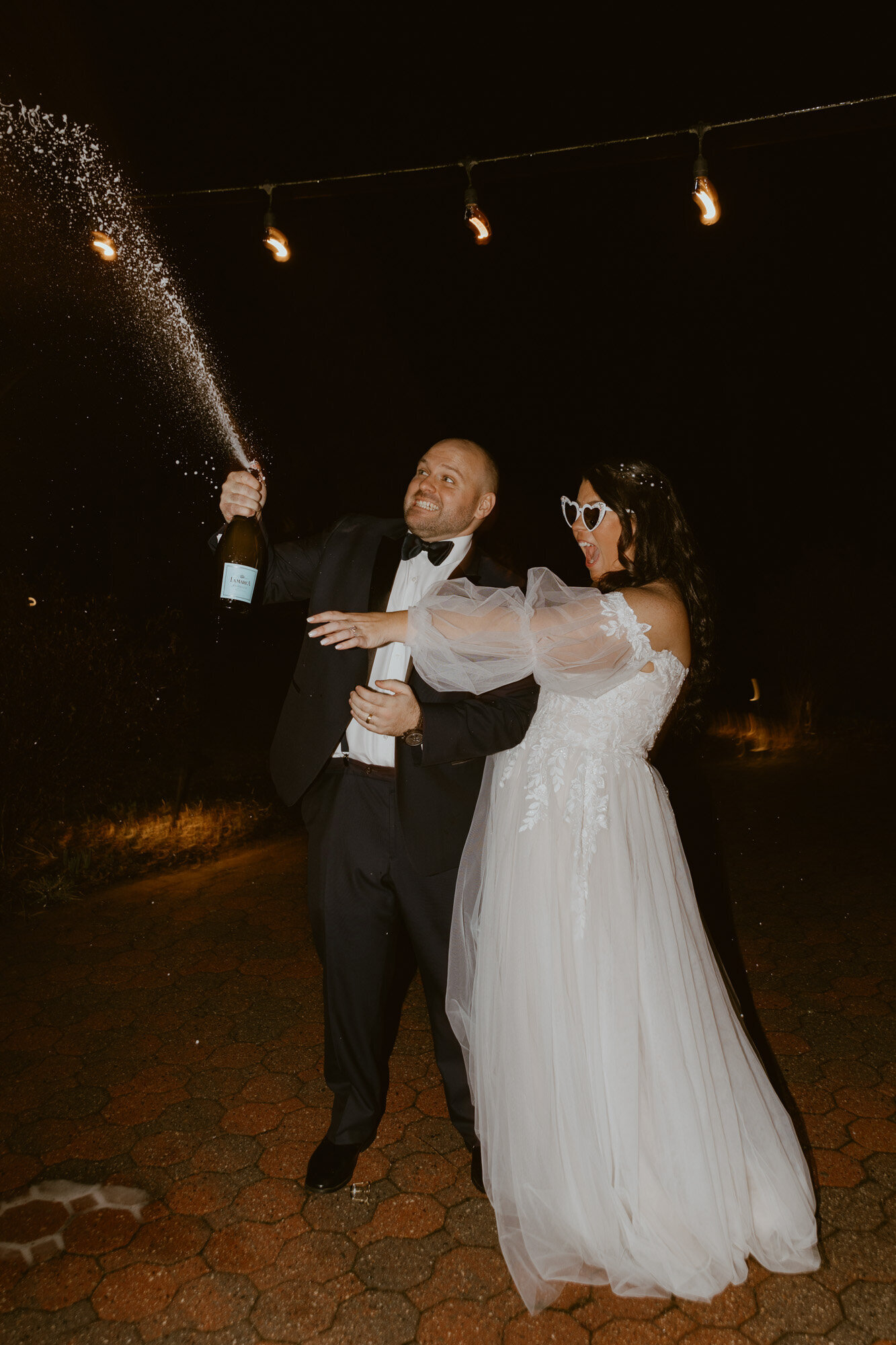 groom spraying a bottle of champagne, and the bride, wearing sunglasses, is holding a drink, smiling