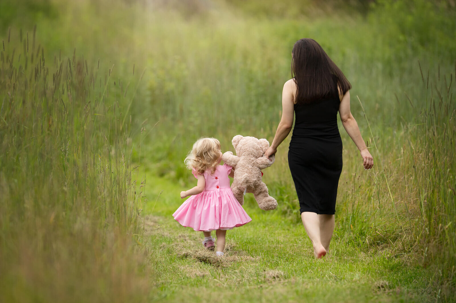 MOther in a black dress walking with two year old daughter in pink dress both holding hands of a teddy bear