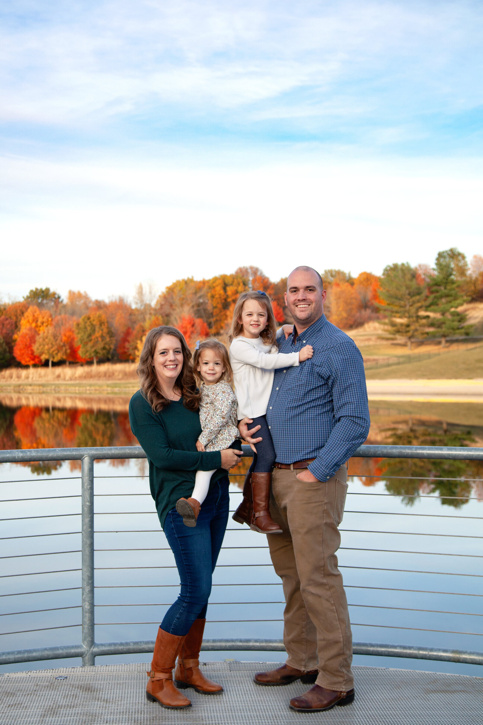 Mom and dad holding their 2 daughters in front of lake and fall trees.