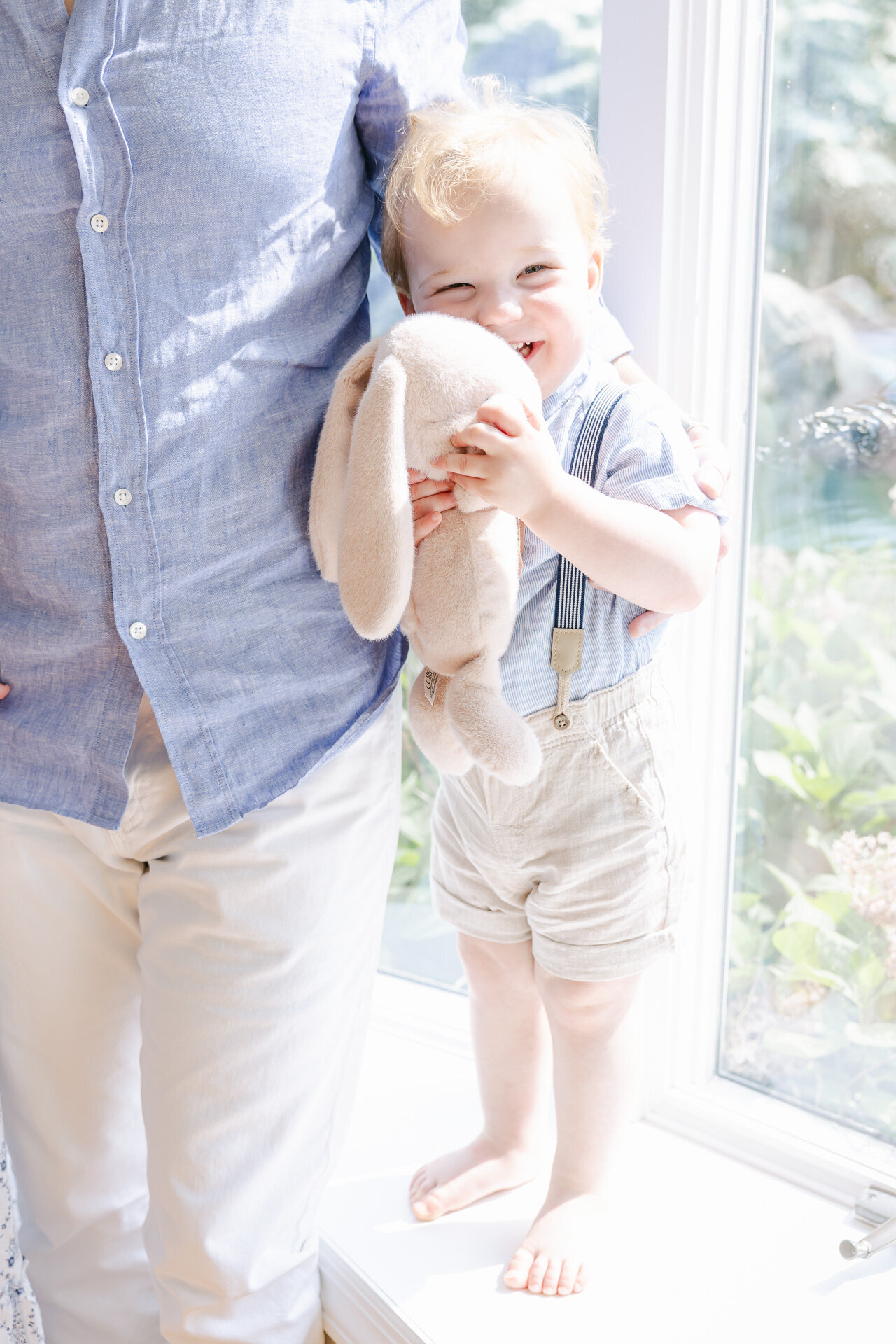A smiling toddler holding a plush bunny toy while standing beside a parent during a sunlit in-home family photography session in Westchester County, captured by Connecticut family photographer Nicole Neira.
