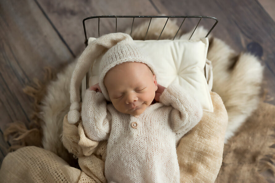 Newborn baby sleeps with his hands behind his head in a little bed.