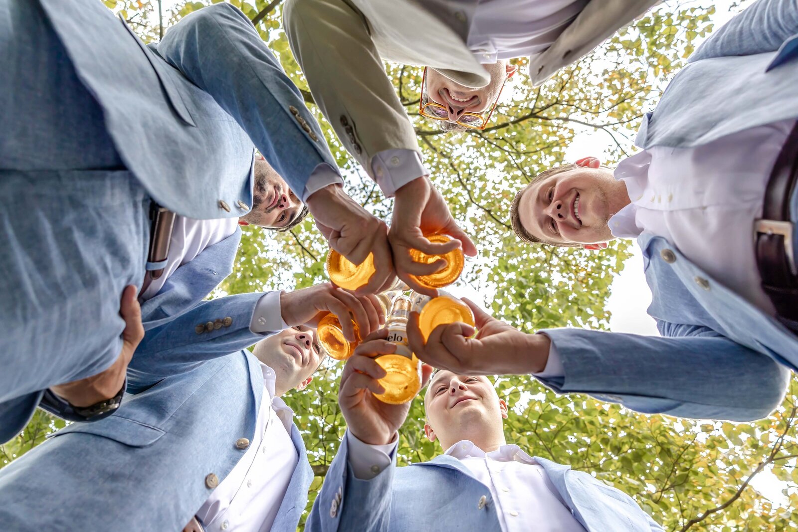 A creative low-angle shot of groomsmen toasting with bourbon glasses under a canopy of trees, captured during a rustic wedding in Boone, NC, blending camaraderie with elegant wedding details.