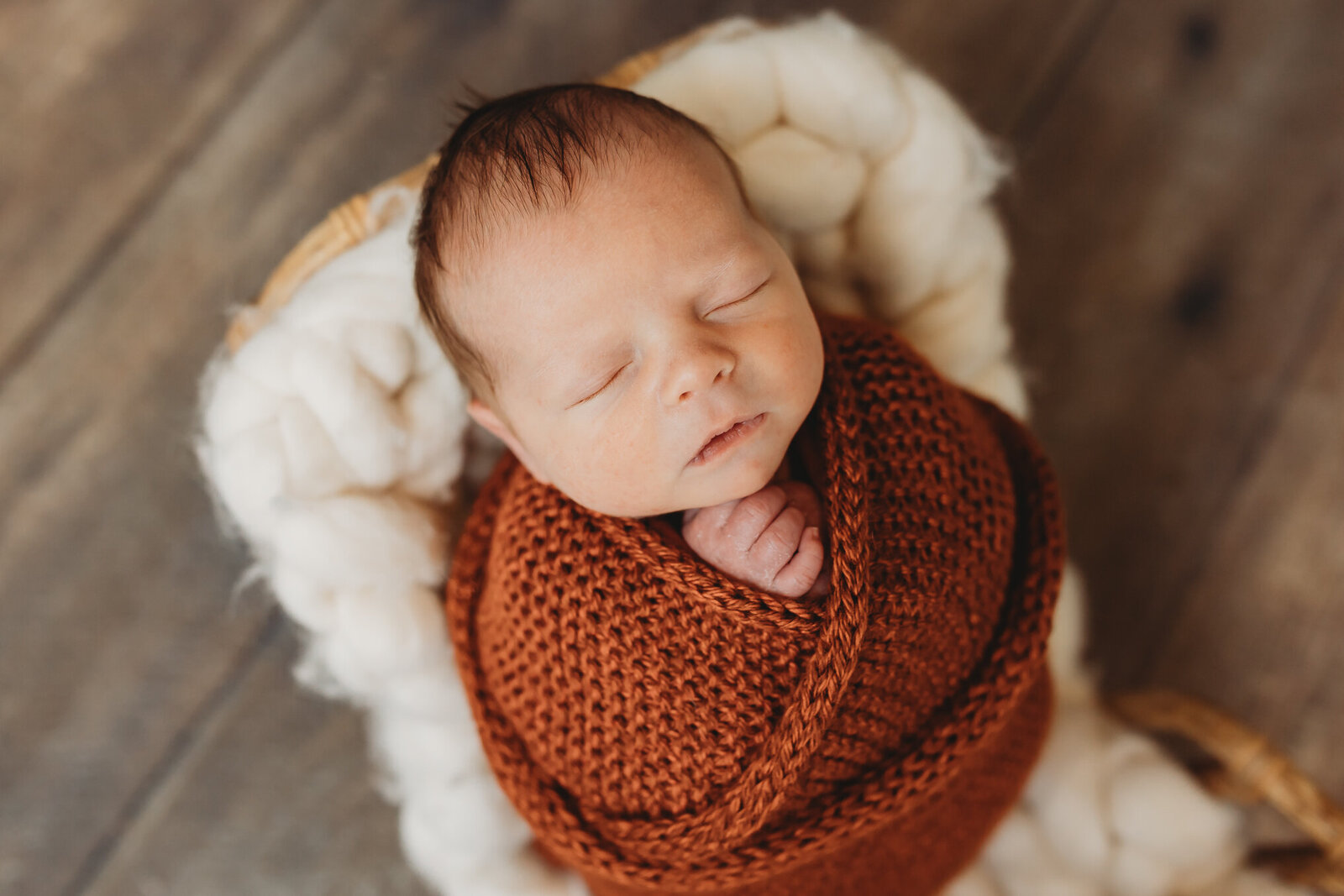 Baby boy swaddled in a burnt orange knit on a bed and wooden floor for newborn photos sleeping