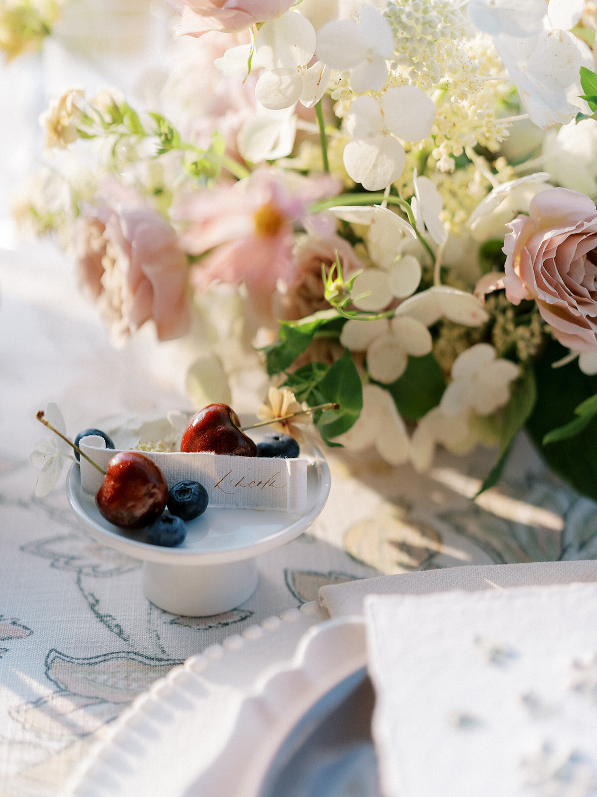 Close up of compote arrangement with taupe and blush garden roses and white hydrangea and table settings accented by cherries and blue berries.