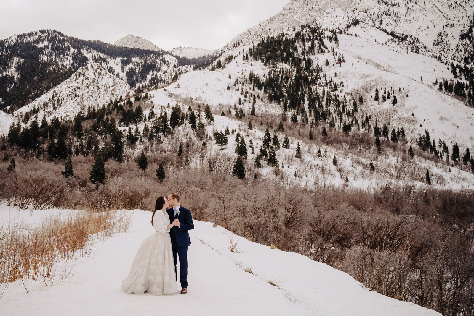 Bride and groom kiss surrounded by a snowy mountain backdrop filled with pine trees. Photo taken by Orlando Wedding Photographer Four Loves Photo and Film.
