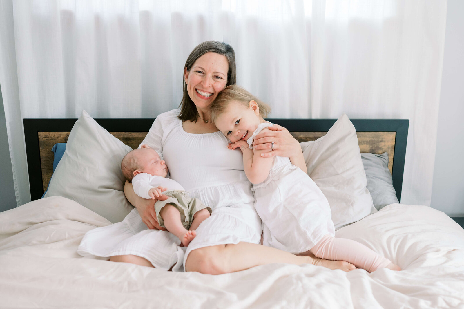 A brunette mom dressed in white sits on her bed and hugs her blonde toddler and her new baby