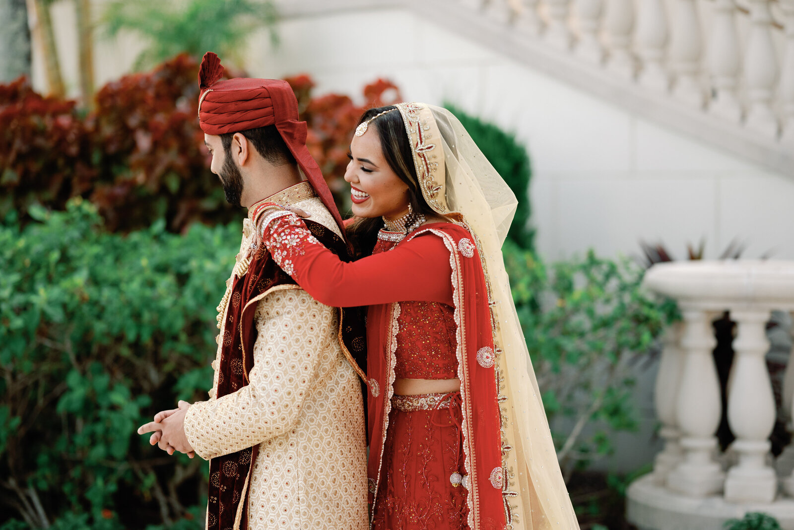 Indian bride hugging her groom from behind at the beginning of their wedding day first look.  She is wearing a traditional red sari.