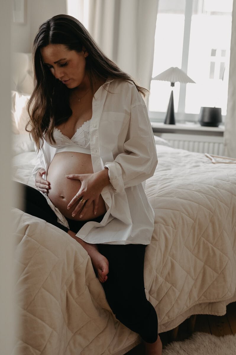 Blivande mamma håller sin mage i händerna sittandes på sängen i sovrummet.