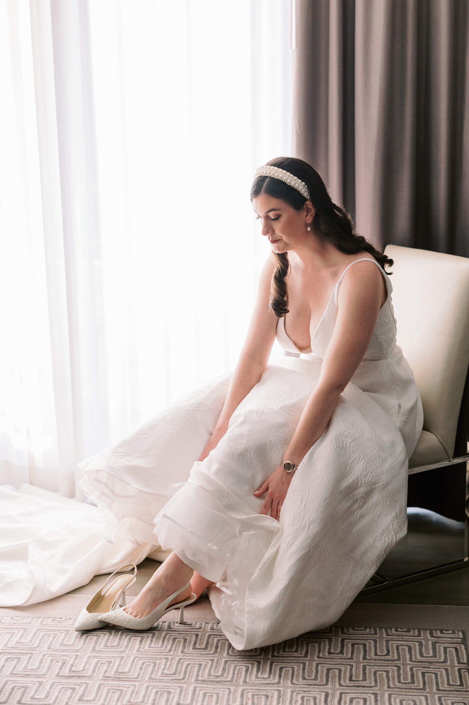 Bride in white dress sits in front of a mirror and puts on her white strappy heeled shoes before her virginia wedding