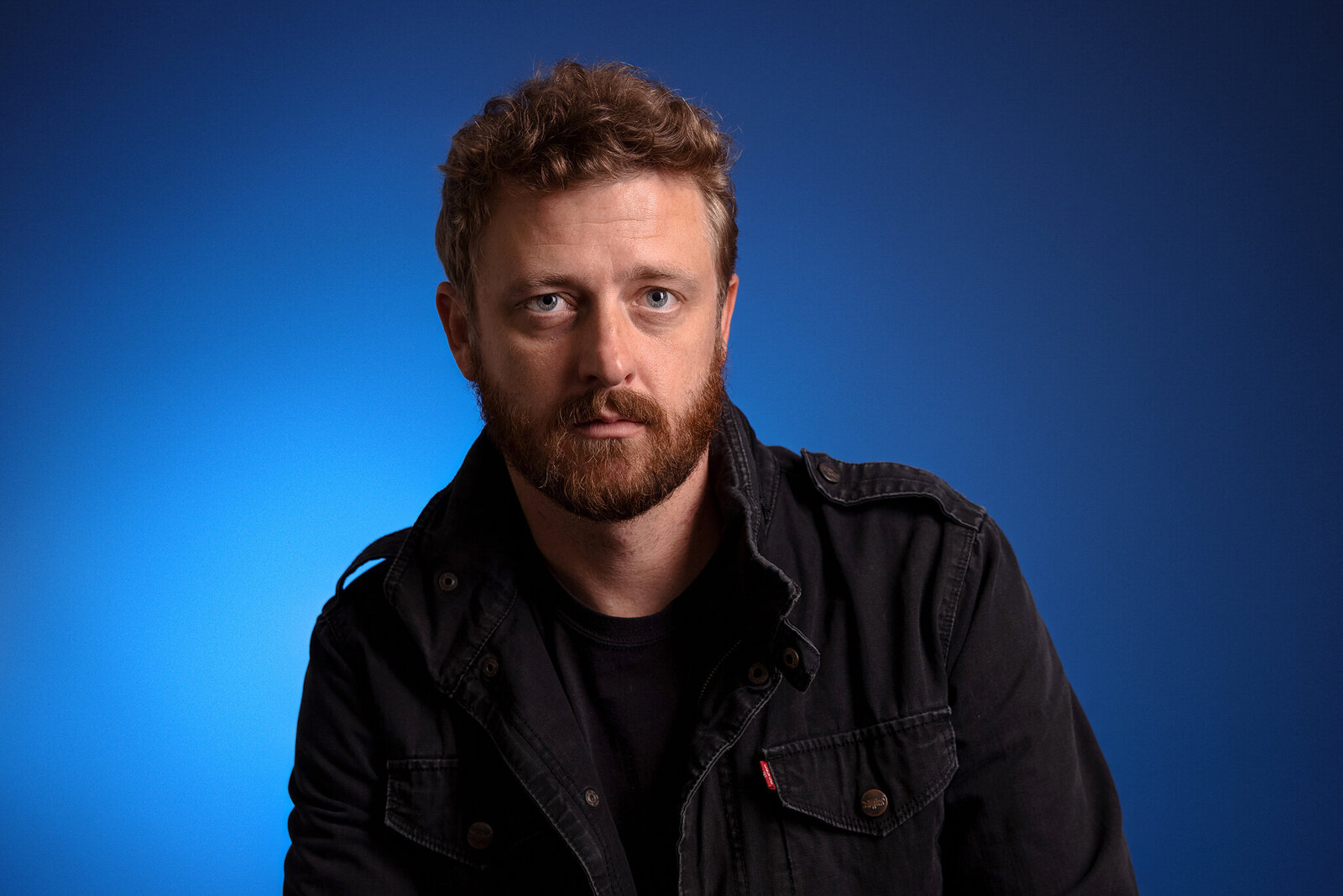 David Benfield with curly light brown hair and a beard wears a black jacket over a black shirt, sitting against a blue gradient background—an example of band photography Jacksonville artists trust for authentic, professional portraits. David Benfield, First Coast News.