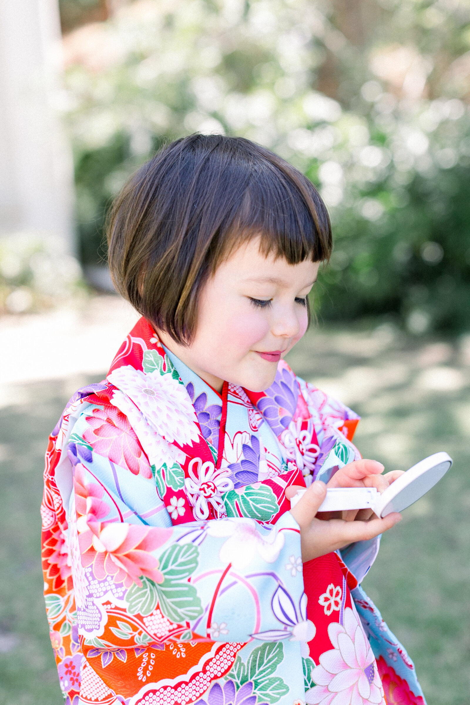 Young girl in vibrant kimono holding a compact mirror during Shichi-Go-San garden photoshoot in Los Angeles | 七五三 ロサンゼルス 庭園でコンパクトミラーを持つ着物姿の女の子