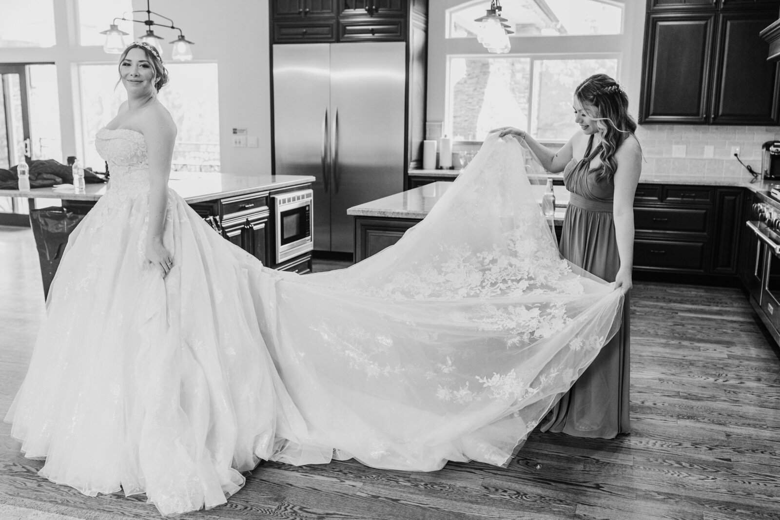 Wolfe Heights wedding pictures with bride standing in a kitchen with maid of honor holding the brides gown and spreading out the train captured by sacramento wedding photographer