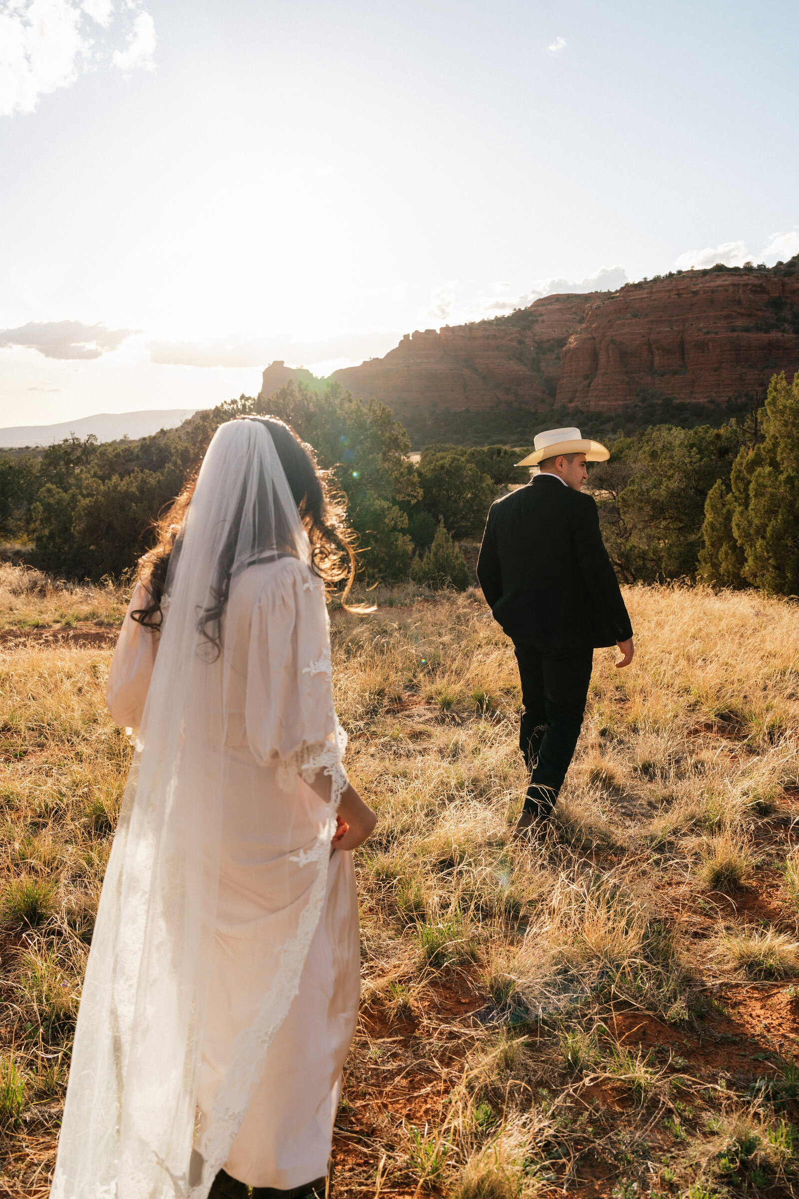 Bride in a vintage lace-trimmed gown and veil follows her groom across the red rock terrain of Bear Mountain at sunset during an adventurous Sedona elopement.