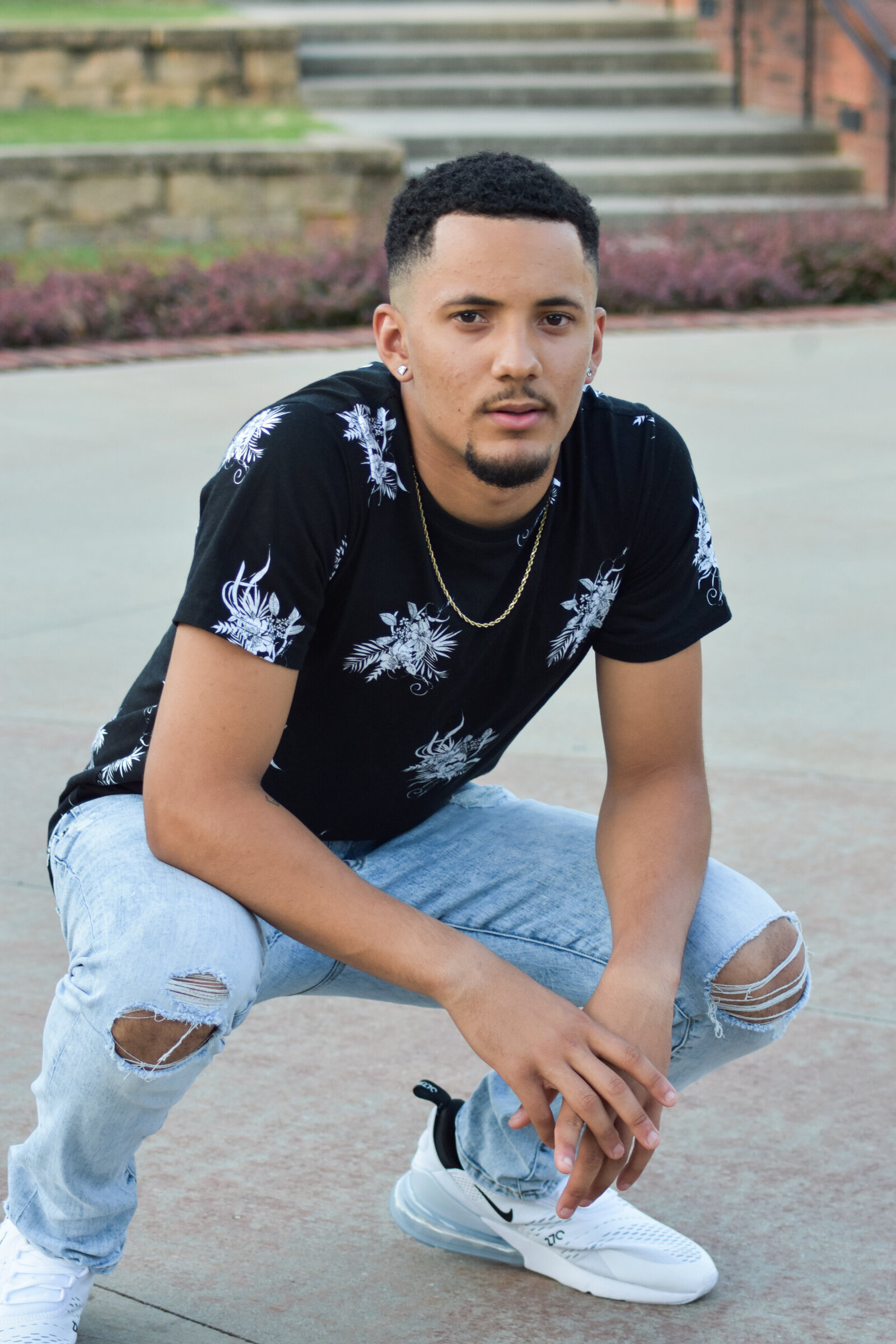 a senior in a black shirt with white flowers crouching down in front of stairs as he poses for his graduation photo photographed by Millz Photography in Greenville, SC