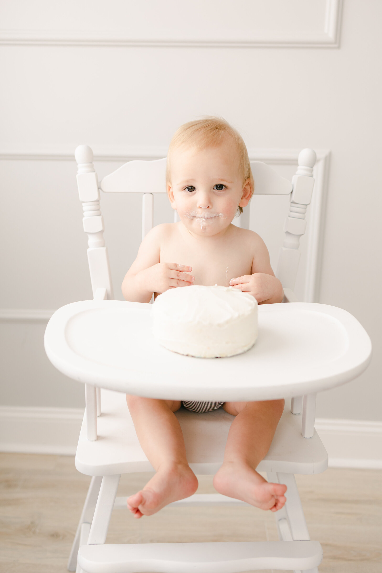 Baby boy sitting in a white high chair with a white cake in front of him waiting to be smashed