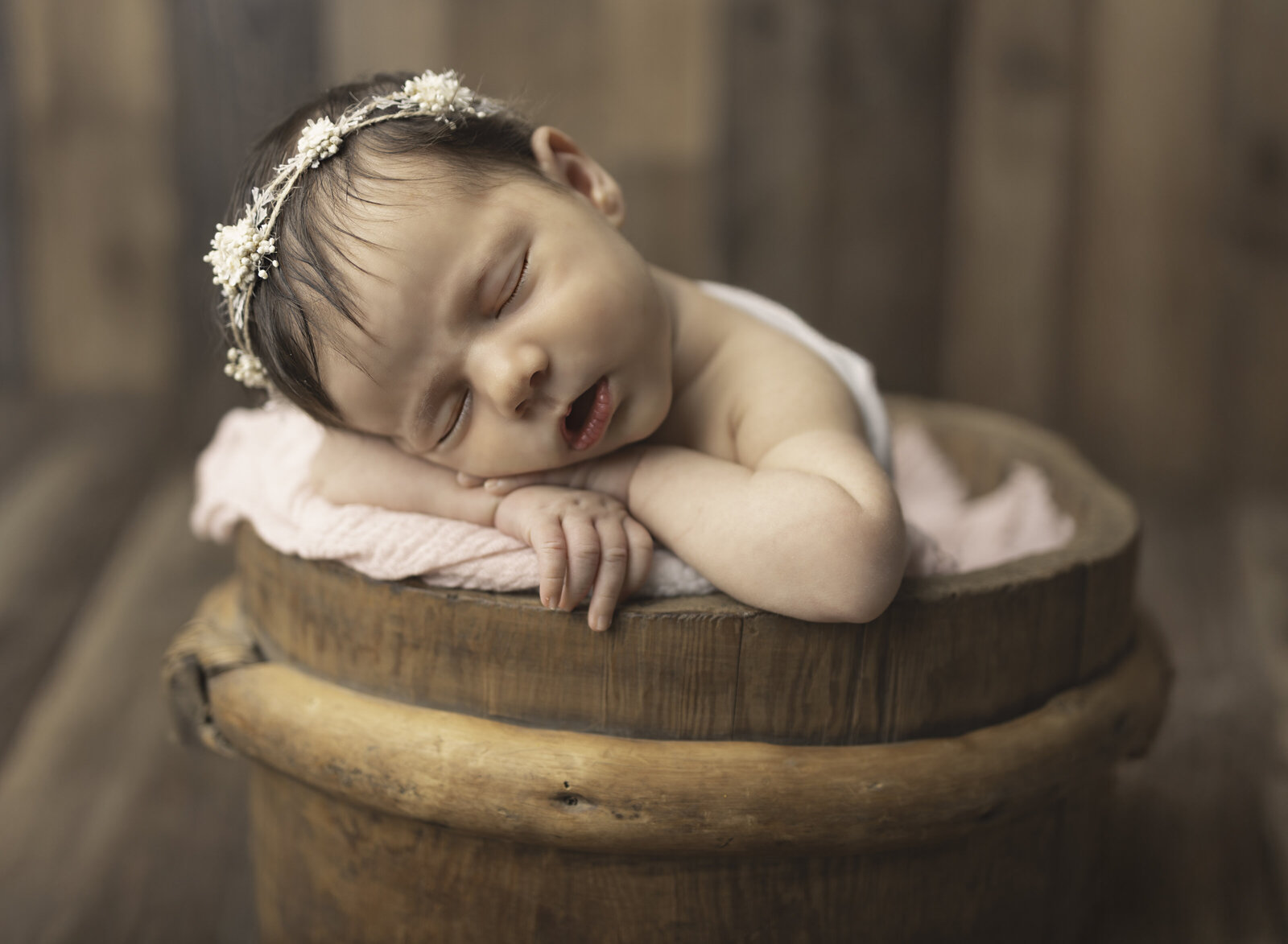 Newborn baby girl wearing floral headband sleeping with arms draped over wooden bucket during NJ photoshoot