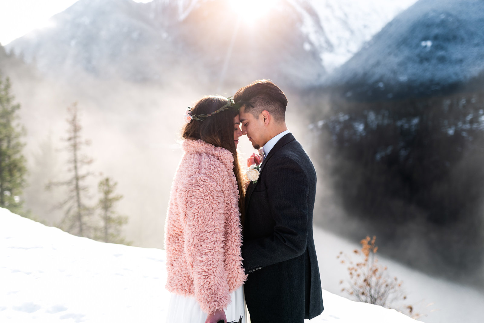 Couple standing on snowy mountain in wedding attire, with foreheads touching and eyes closed, with smiles on their face.