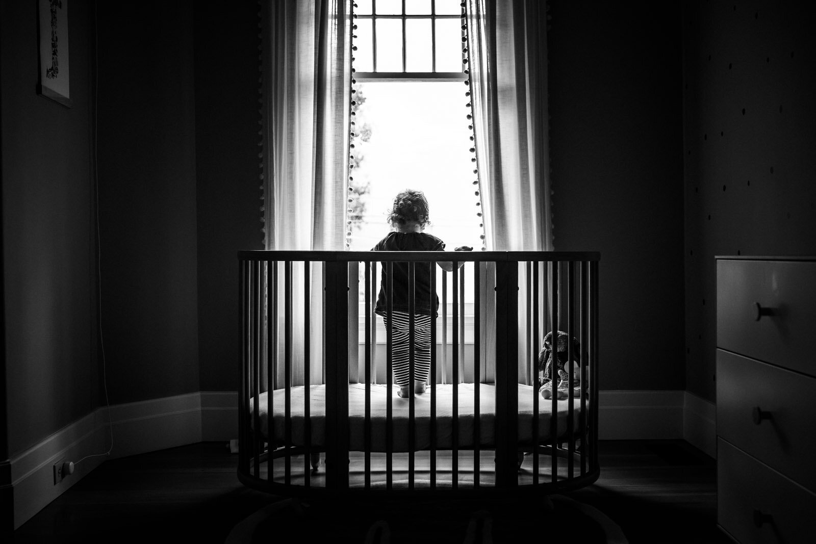 little girl standing in crib and looking out the window