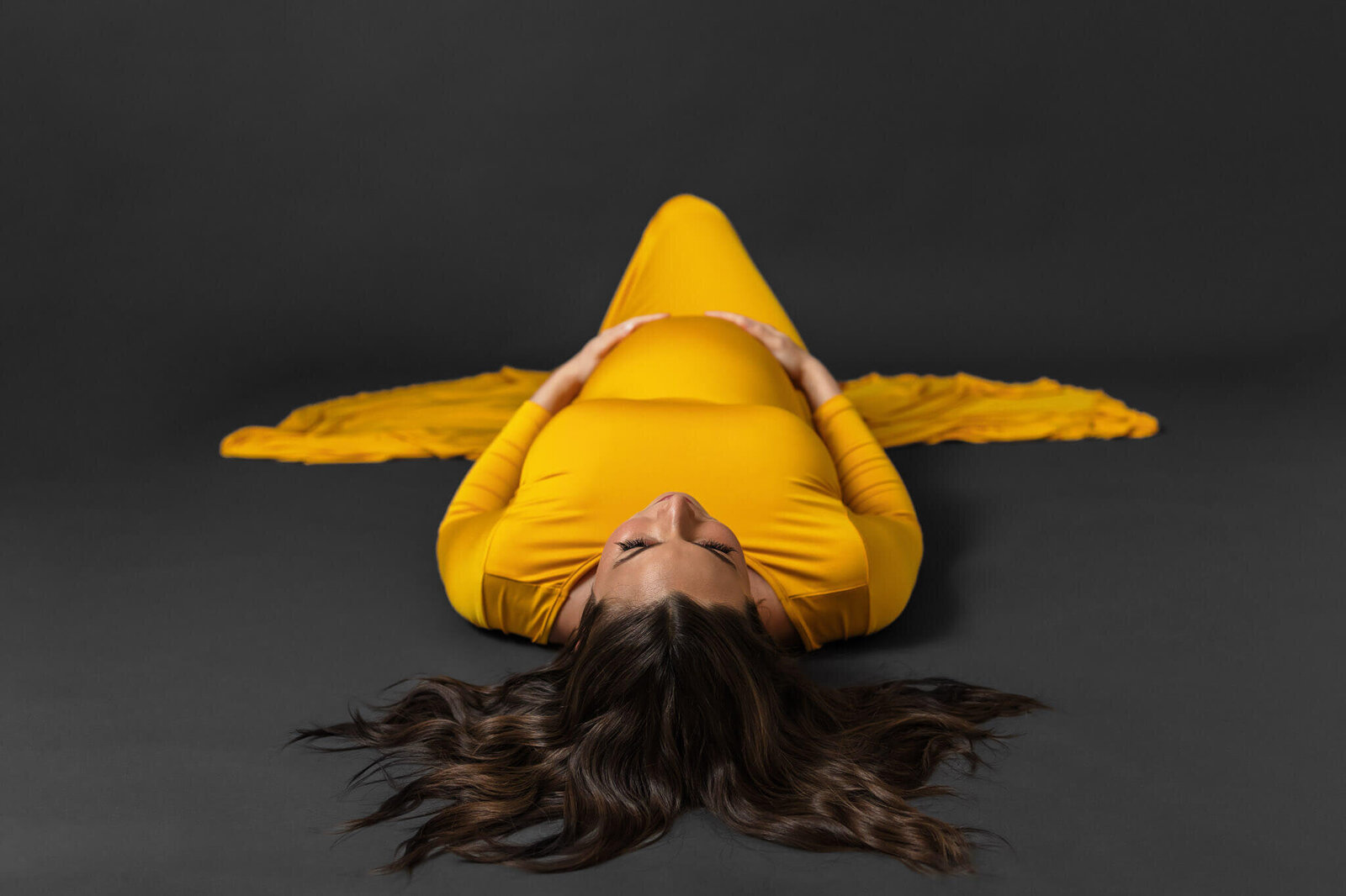 An expecting woman in a yellow dress laying on the ground of the studio hugging her belly.