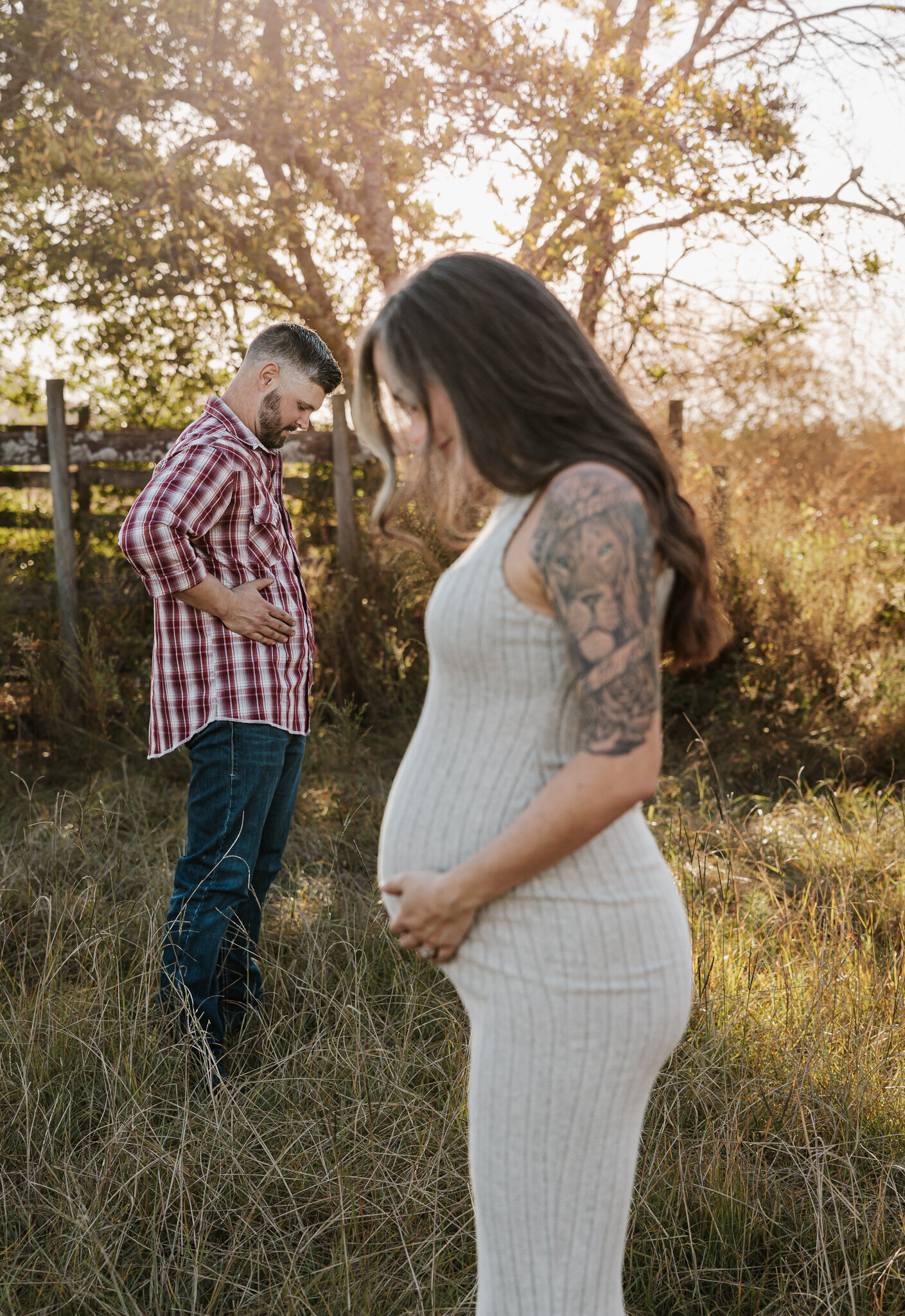 Outdoor maternity session in Aiken SC - expecting mother in a ribbed dress holding her belly  while the father stands behind her in a golden field playfully holding his belly.