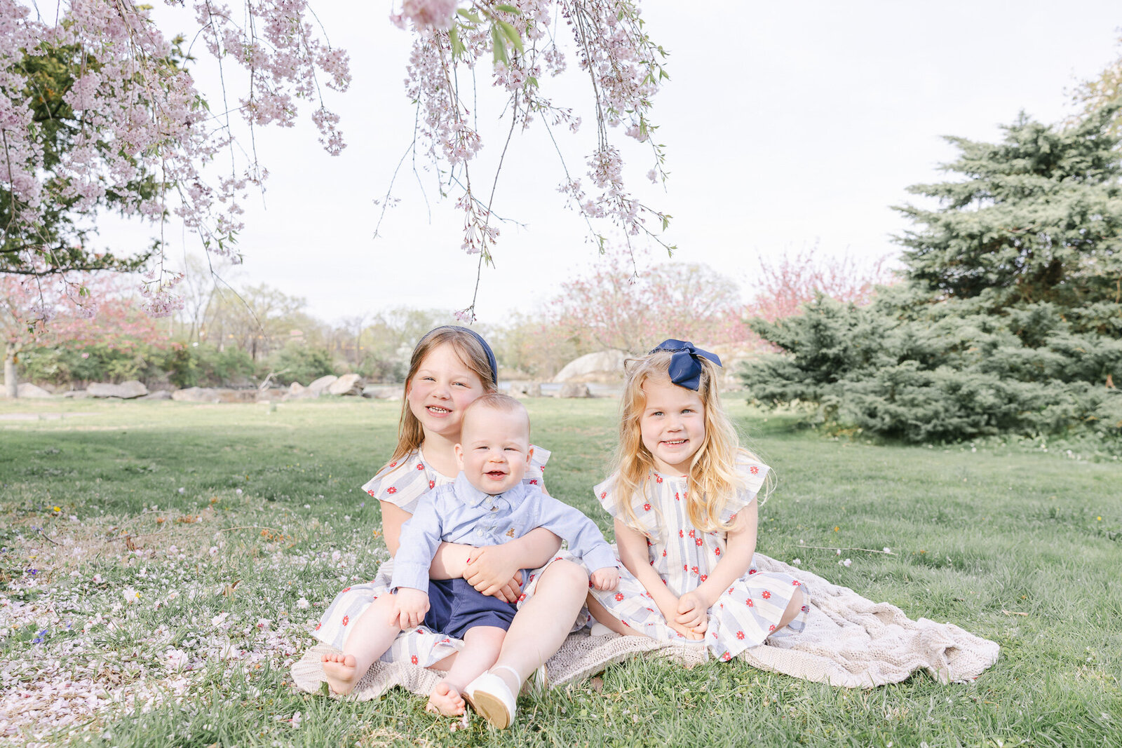 Three young siblings sit on a blanket beneath blooming cherry blossom branches. Two sisters in matching cherry-print dresses sit beside their baby brother, who wears a blue shirt and navy shorts. Captured on a soft spring day, this sweet sibling portrait is full of joy, connection, and pastel tones—perfect inspiration for seasonal family photos.