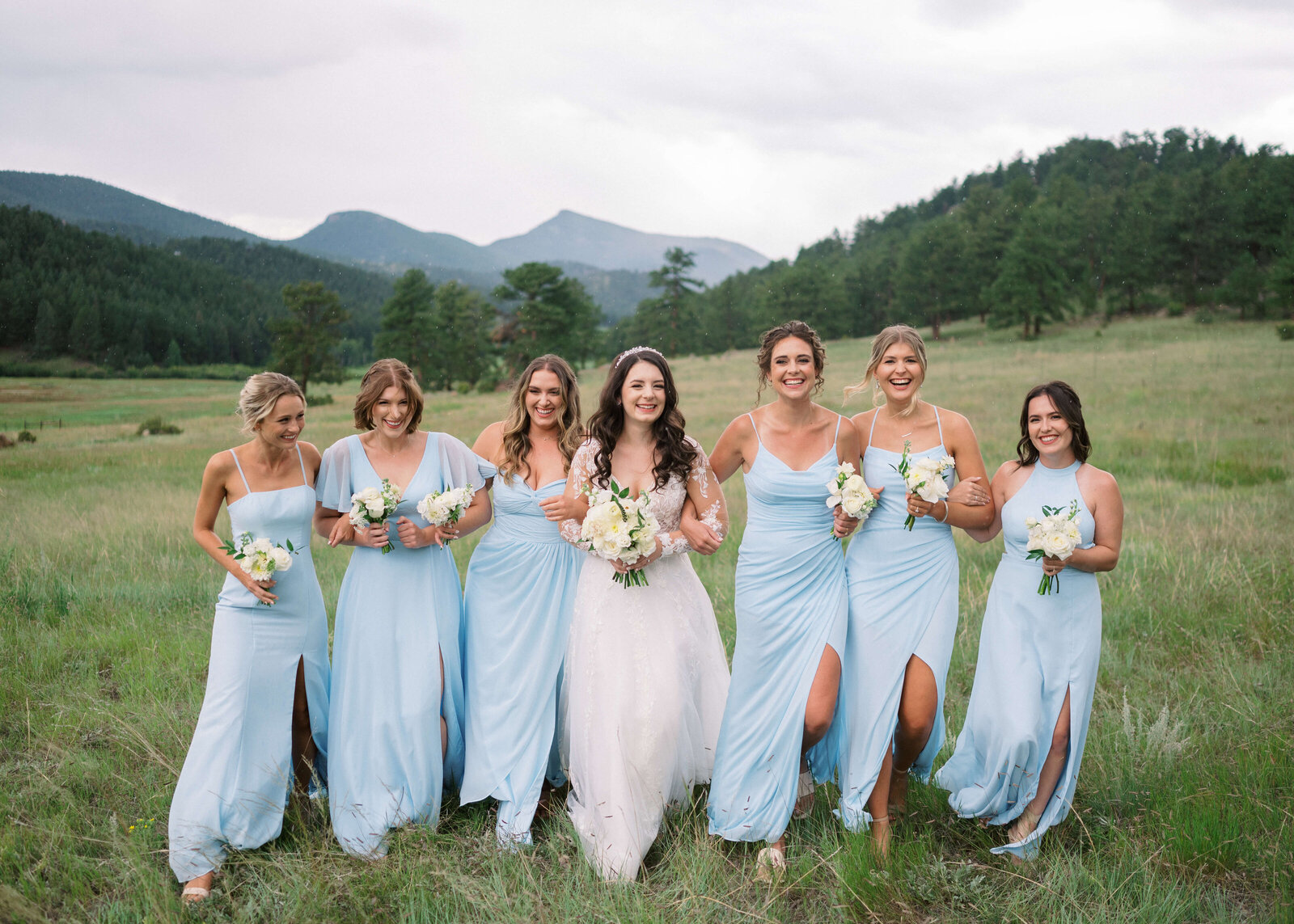 bridal party dressed in light blue bridesmaids dresses walk in a field with their friend and bride