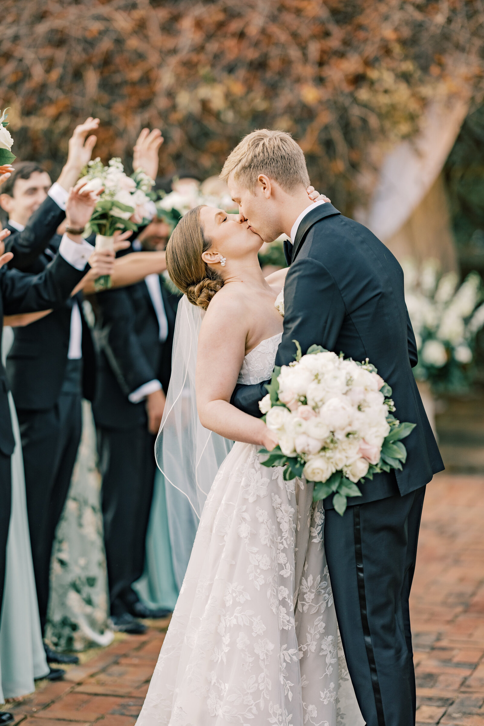 Bride and groom share a romantic kiss after walking through a tunnel of love created by their bridal party, celebrating their union with cheers and excitement.