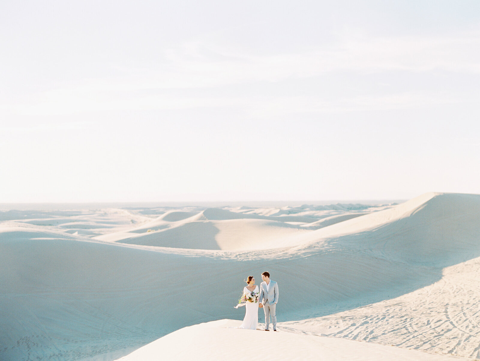 Kohnur epic couple portrait with bride in natural minimal makeup and sleek low updo wearing a flowy gown on vast white sand dunes.