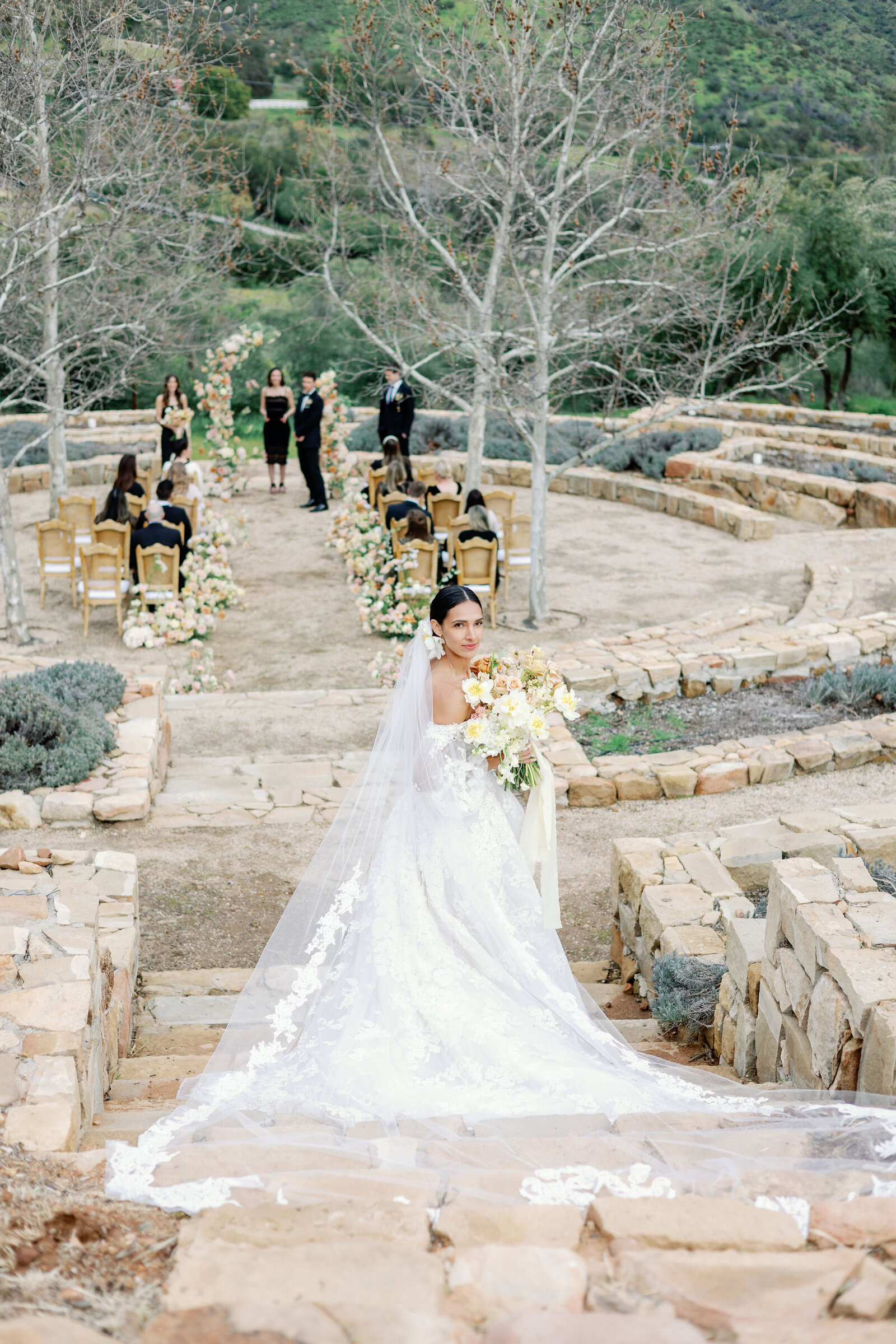 bridal portrait in outdoor wedding venue ceremony space with bride walking down a stone stair way towards the ceremony while looking back over her shoulder captured by wedding photographer bay area