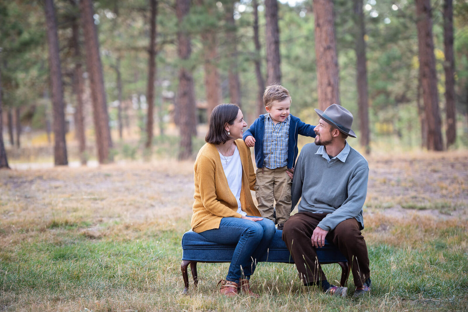 A toddler boy stands in a bench in a park between mom and dad
